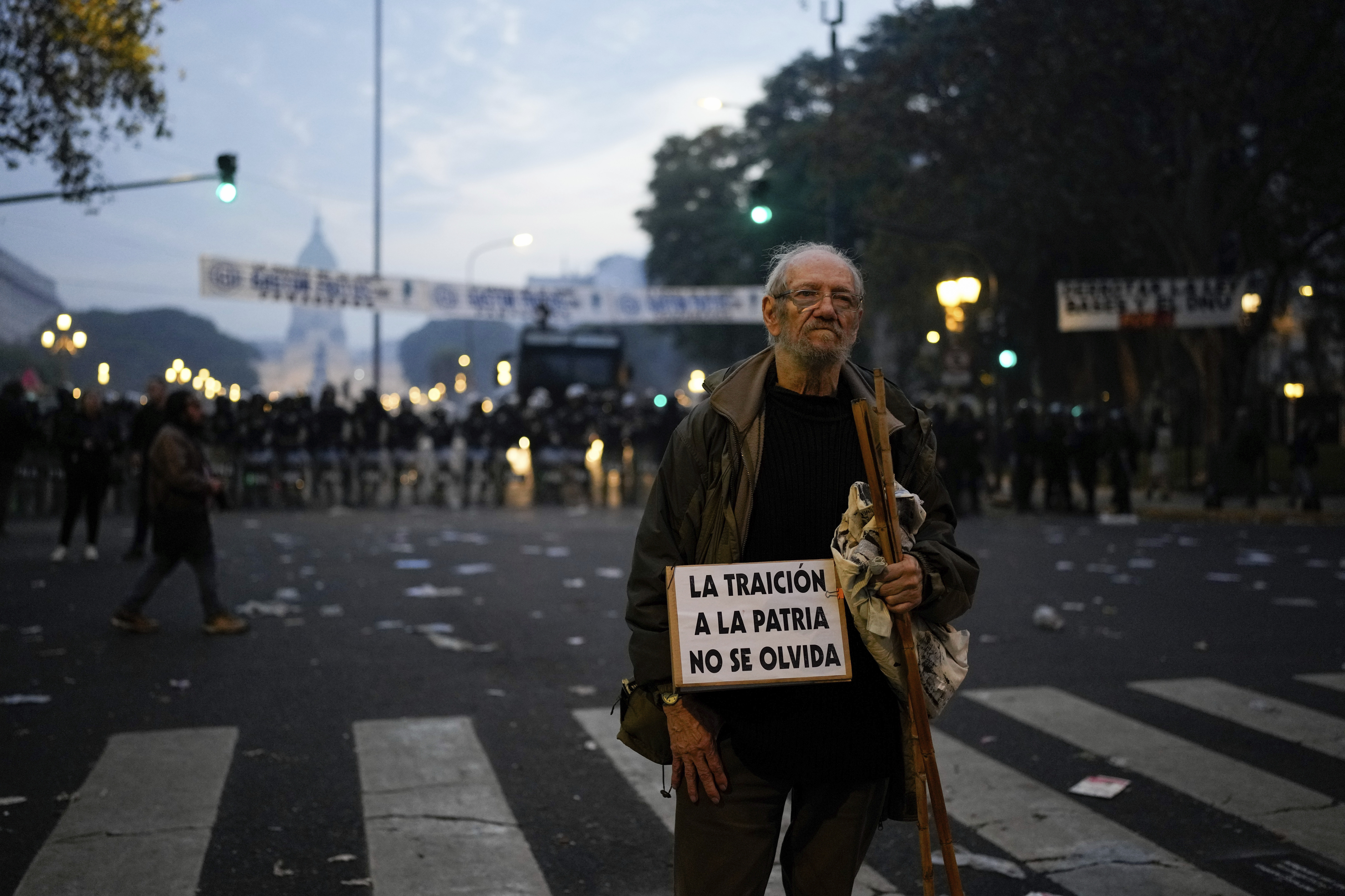 An elderly man holds a protest sign on the road in Argentina. Behind him, the dome of Congress is visible.