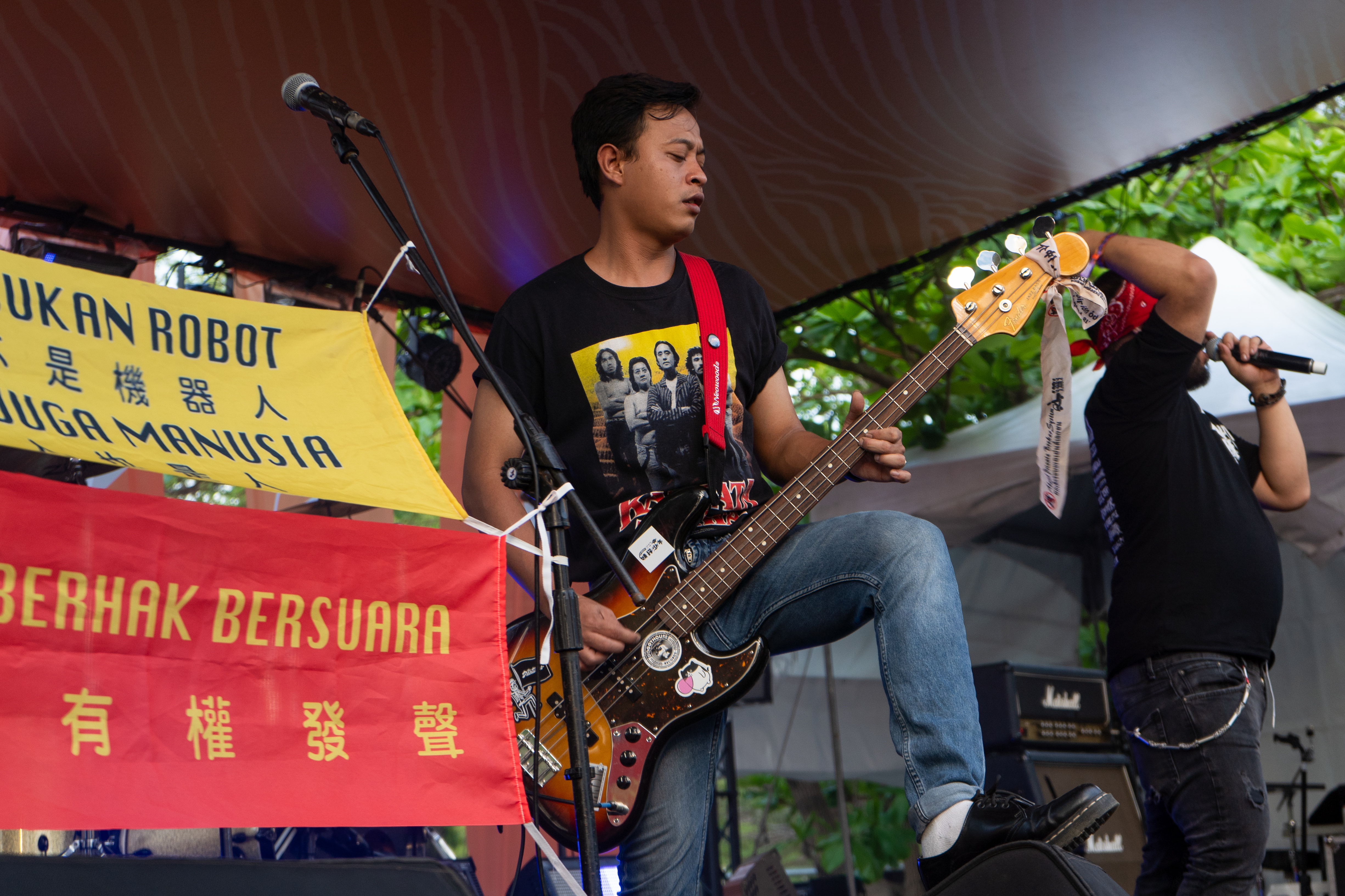 Southern Riot's guitarist playing on stage. There are banners in Indoneesian saying 'We are not robots' and 'We are humans too'. The guitarist has curly black hair and is wearing jeans and a black T-shirt. He has his left foot up on one of the speakers and is balancing the guitar on his knee