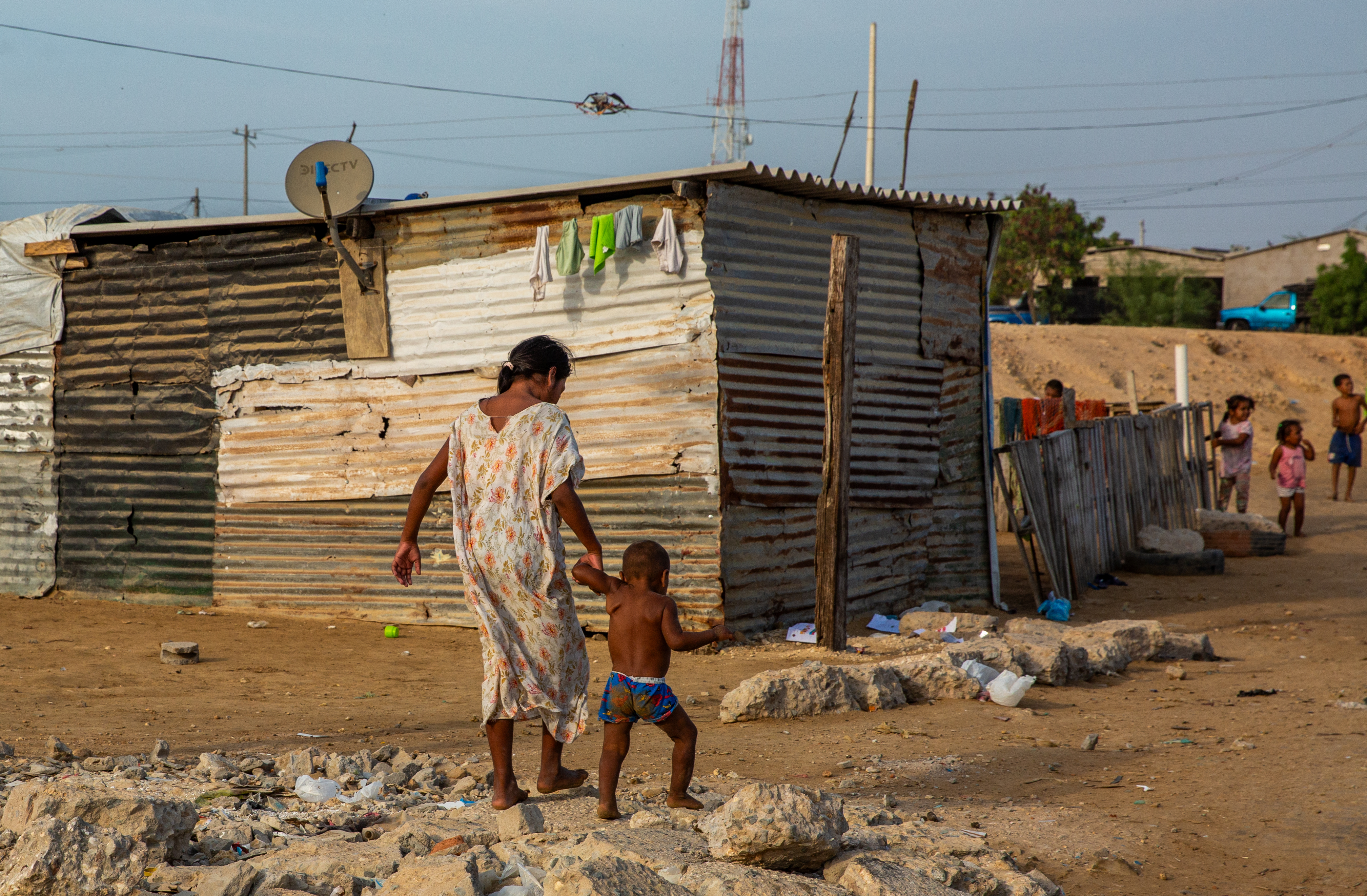 A woman and child walk hand in hand through the drylands of La Guajira, a ramshackle house in the background with laundry hanging under its roof.