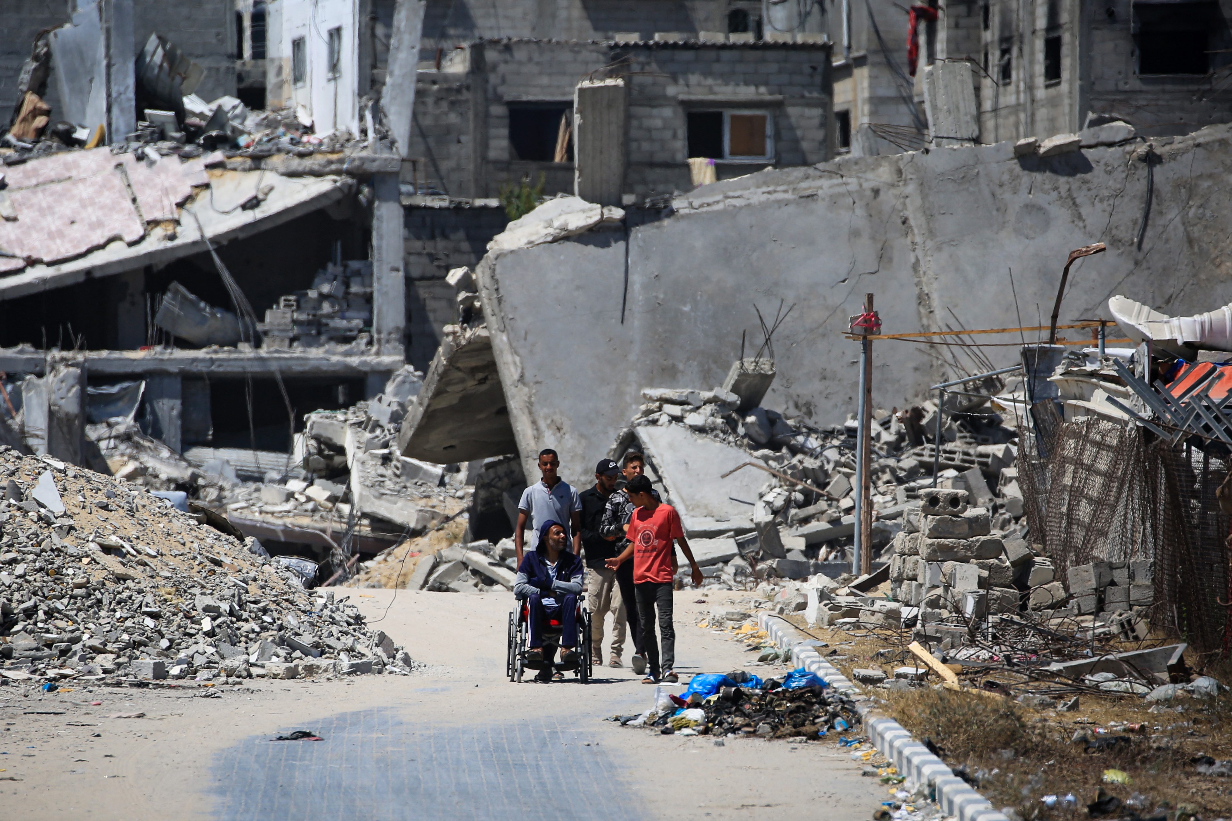 Palestinians walk alongside a man on a wheelchair near buildings destroyed during previous Israeli bombardments, in Khan Yunis in the southern Gaza Strip on July 3, 2024, amid the ongoing conflict between Israel and the Palestinian Hamas militant group. (Photo by Eyad BABA / AFP)