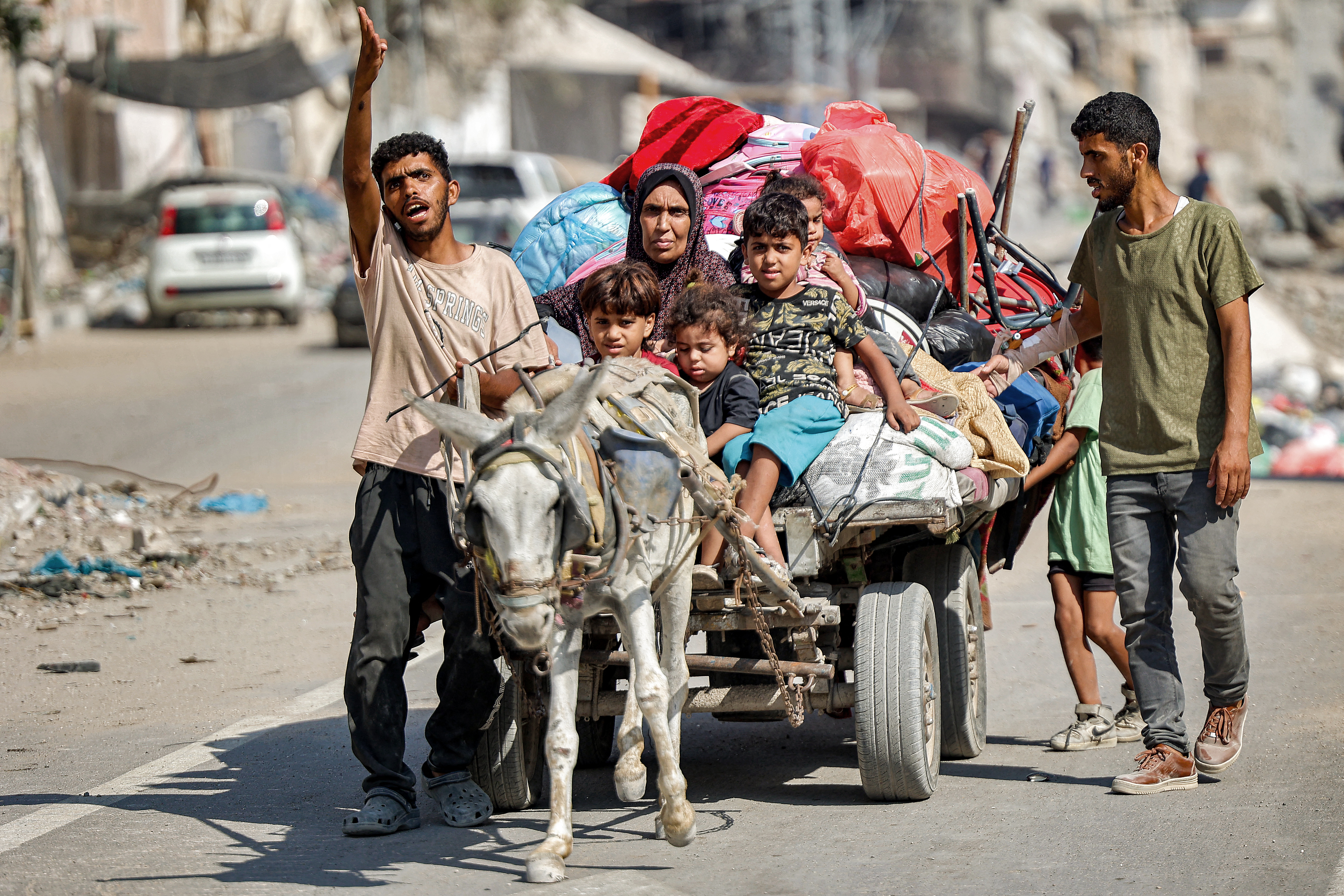 A man gestures while leading a donkey pulling a cart transporting his family members and belongings