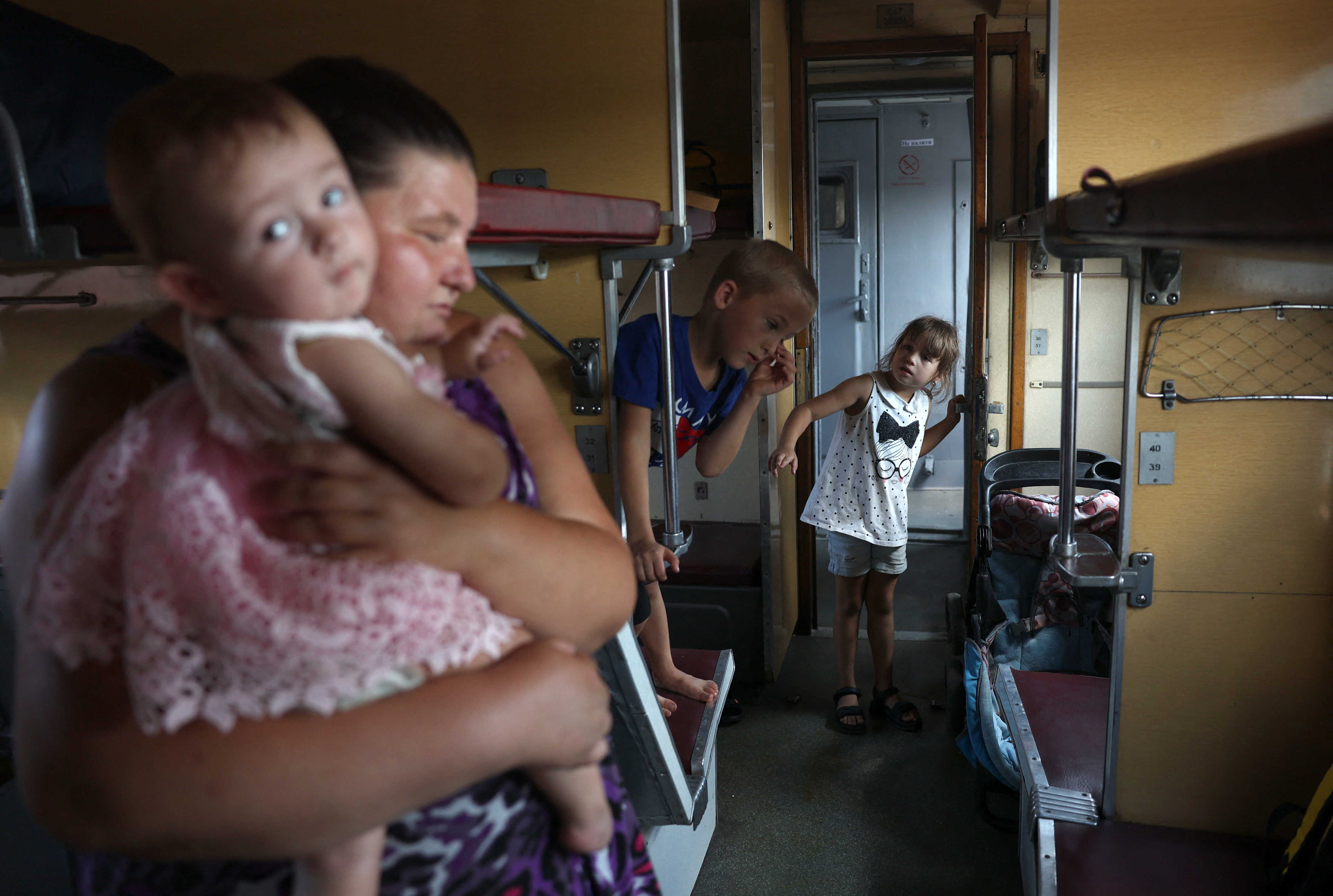 A woman holding a baby in her arms inside a railway carriage. Two young children are also in the carriage. One is sitting on one of the bunks. The baby is looking at the camera.