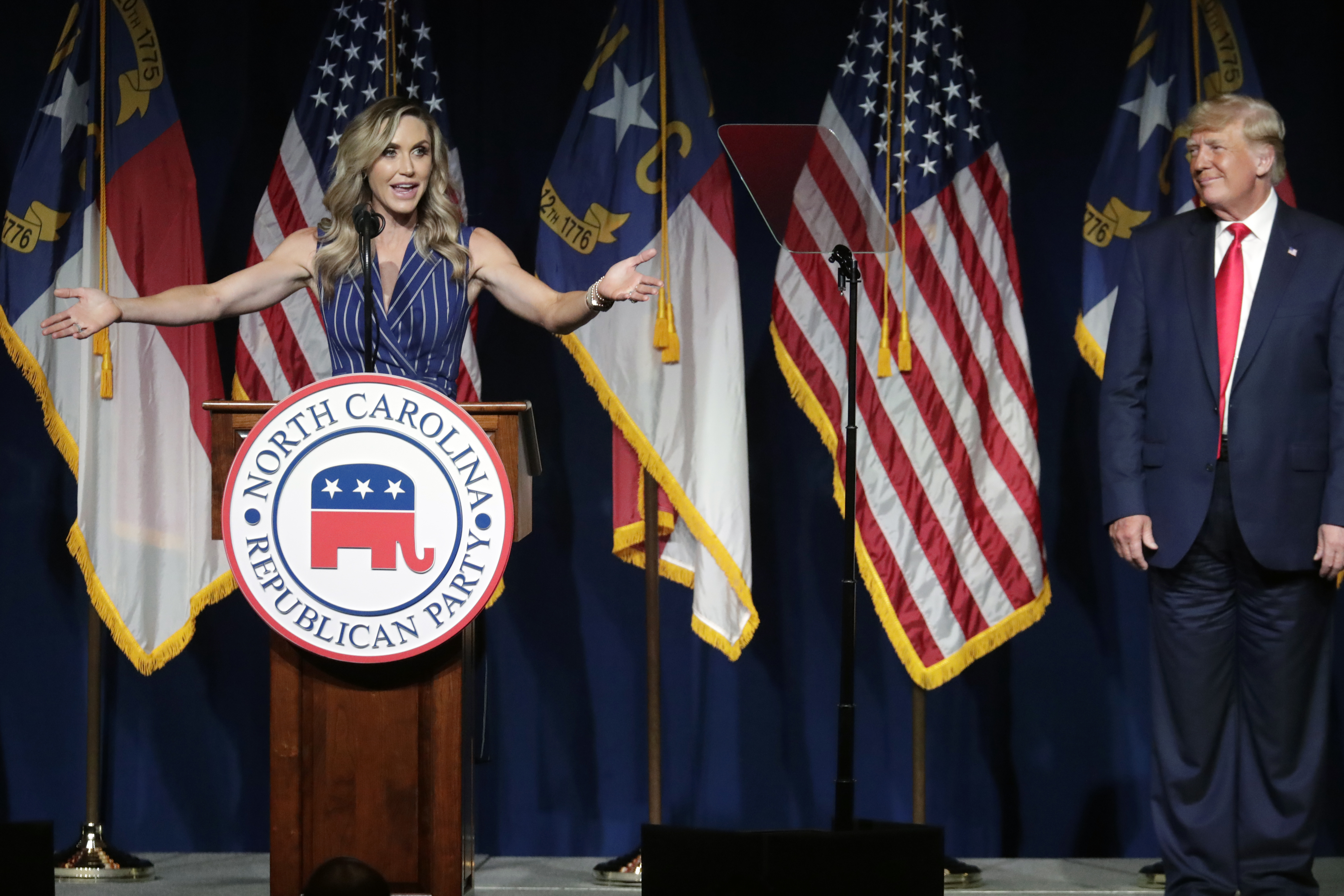 Lara Trump stands at an RNC podium, her arms outstretched, as her father-in-law looks on from another side of the stage.