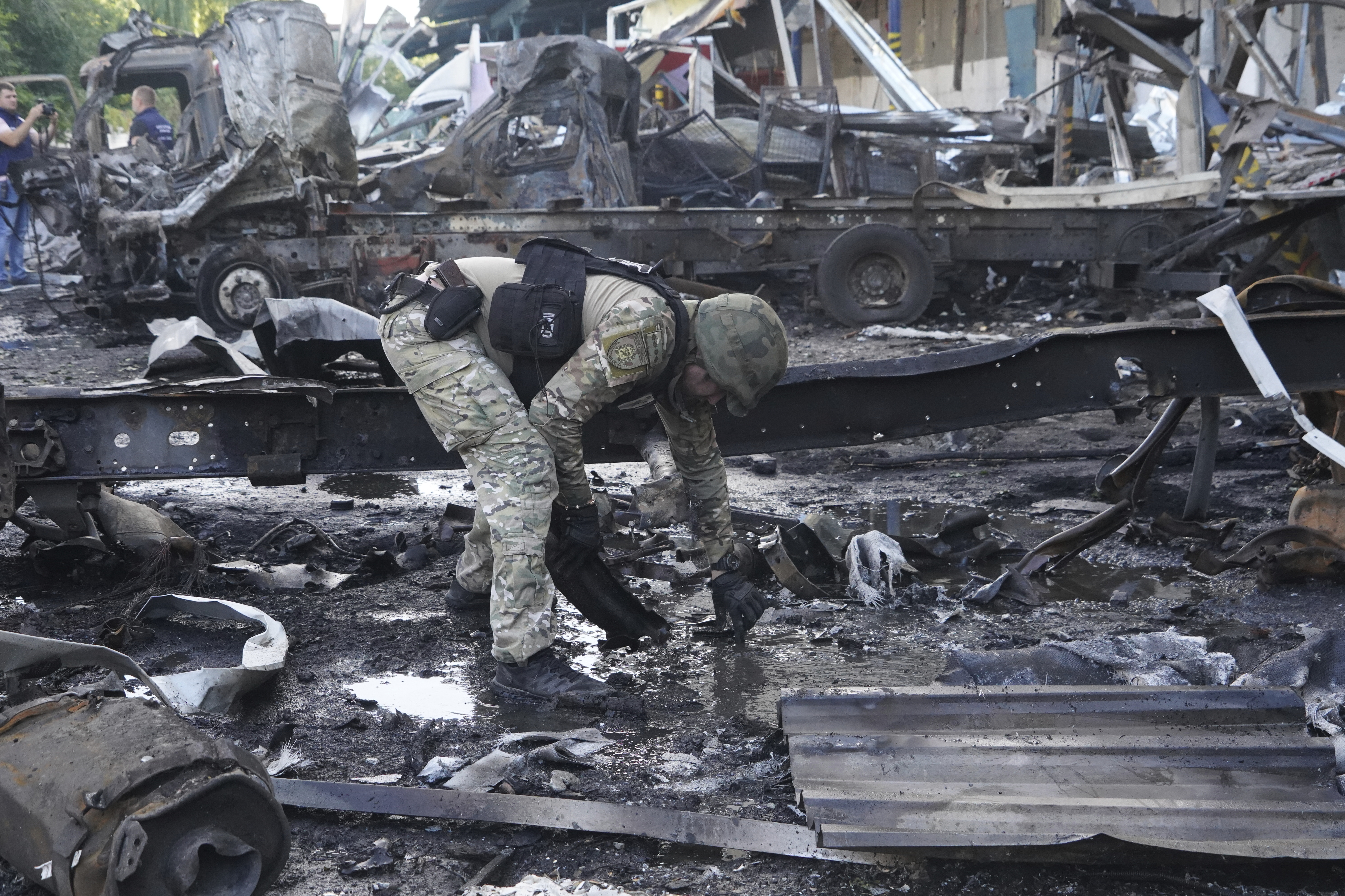 A police officer collecting evidence after the attack on the postal warehouse. He is bending down to pick up something from the ground. There are burned out trucks and vans behind him. Everything is grey and black.