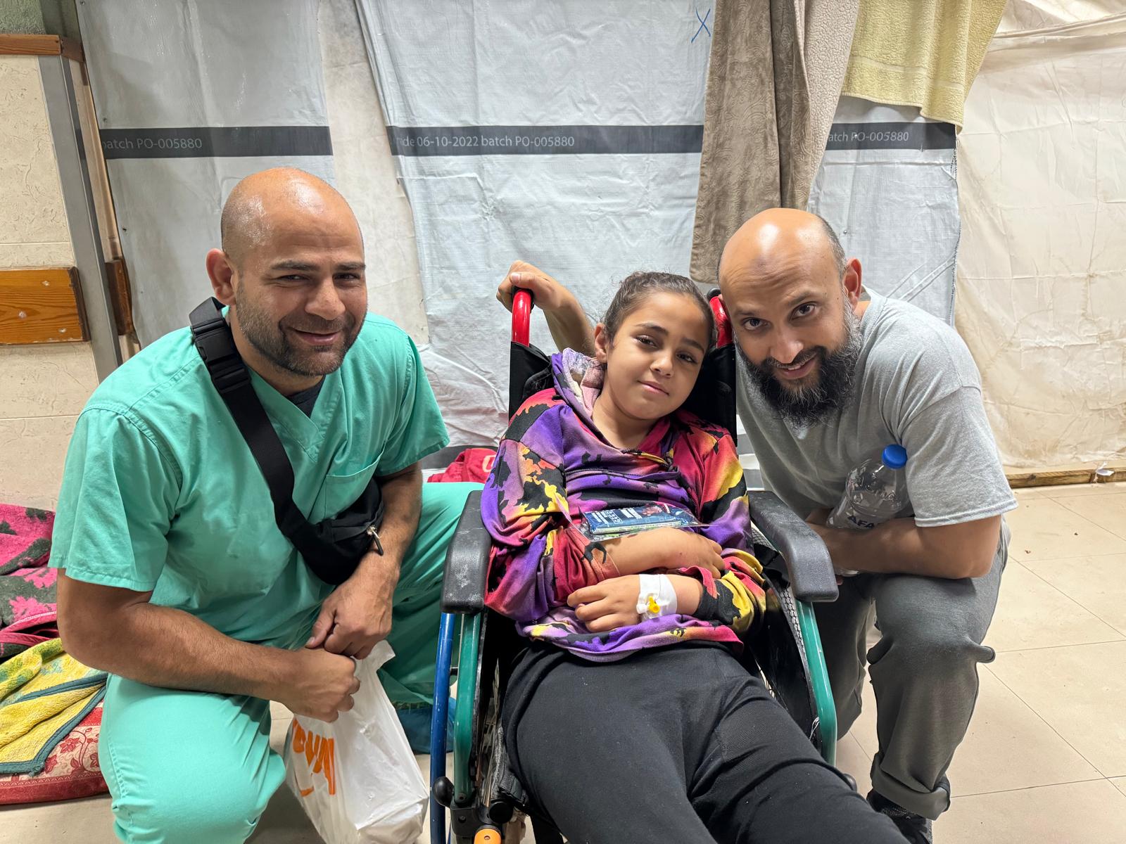 Omar Sabha kneels in teal scrubs next to a young girl in a wheelchair.