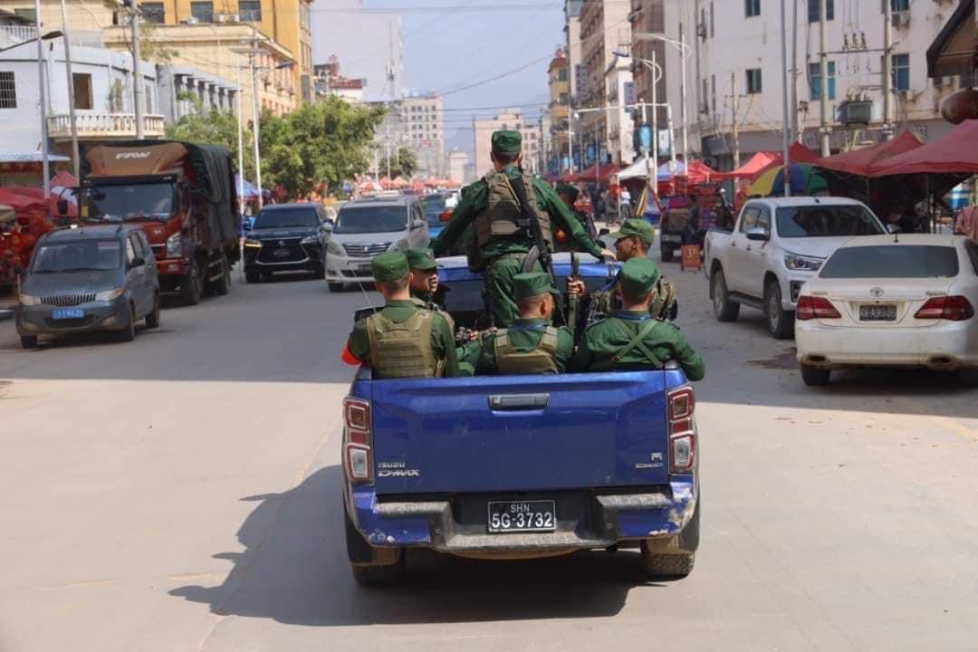 Soldiers on the back of a blue pick-up truck drive along a Laukkai street. There are tall buildings on either side, and other vehicles on the road. They are driving away from the camera.