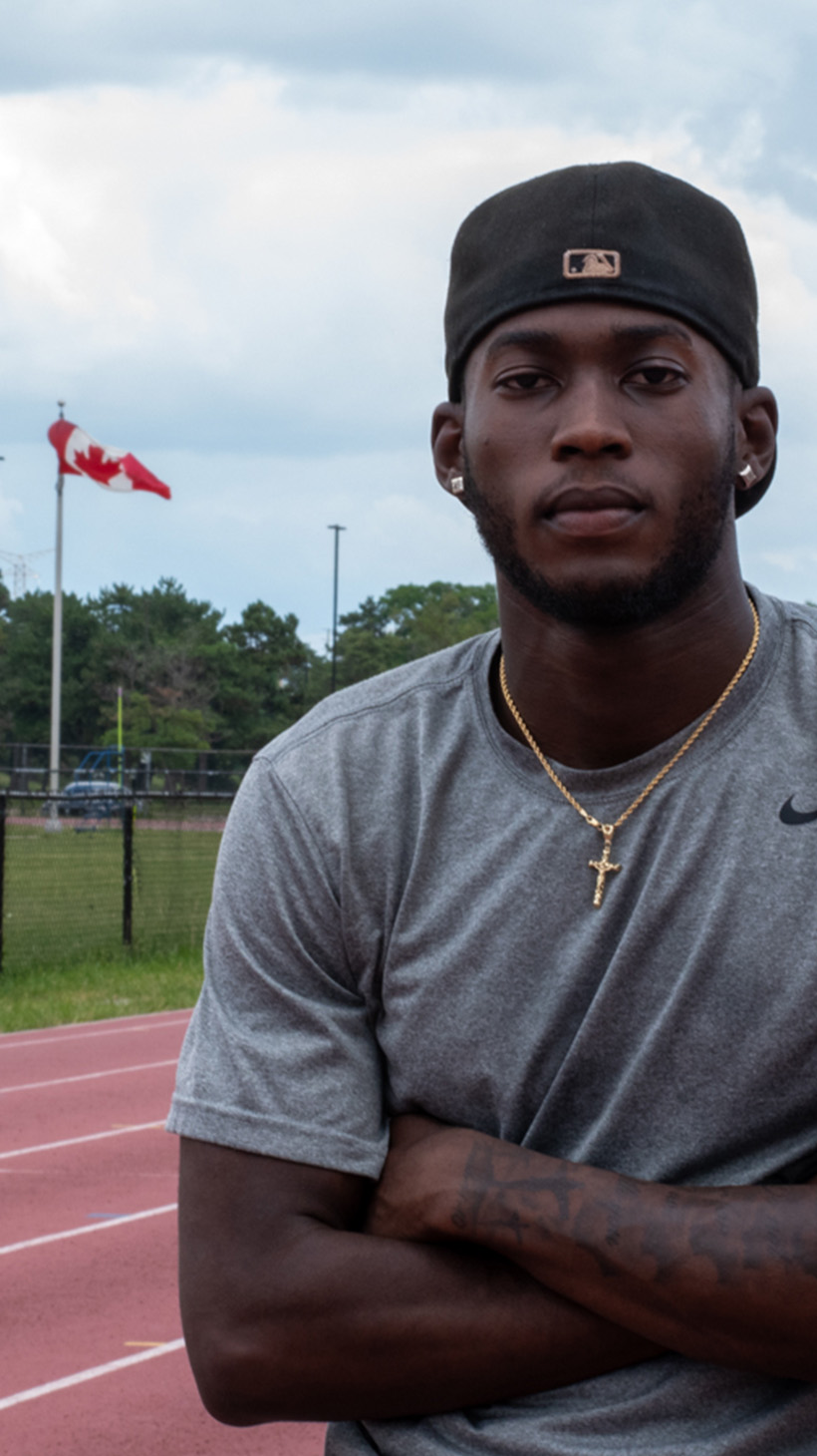 Tamarri stands on an outdoor track cross-armed. Behind him is a flag pole bearing the Canadian flag