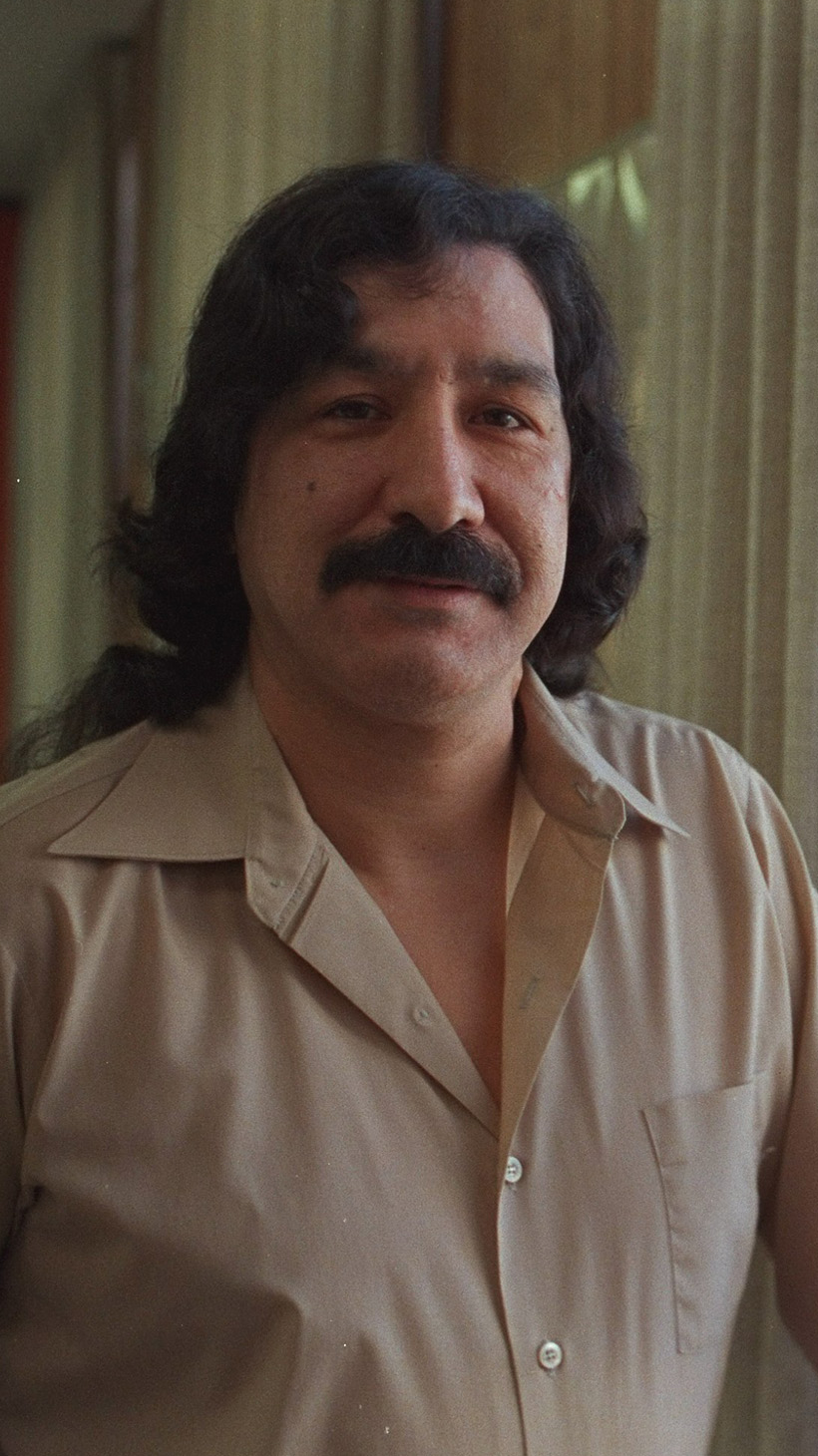 Leonard Peltier stands at a prison window, dressed in a beige collared shirt.