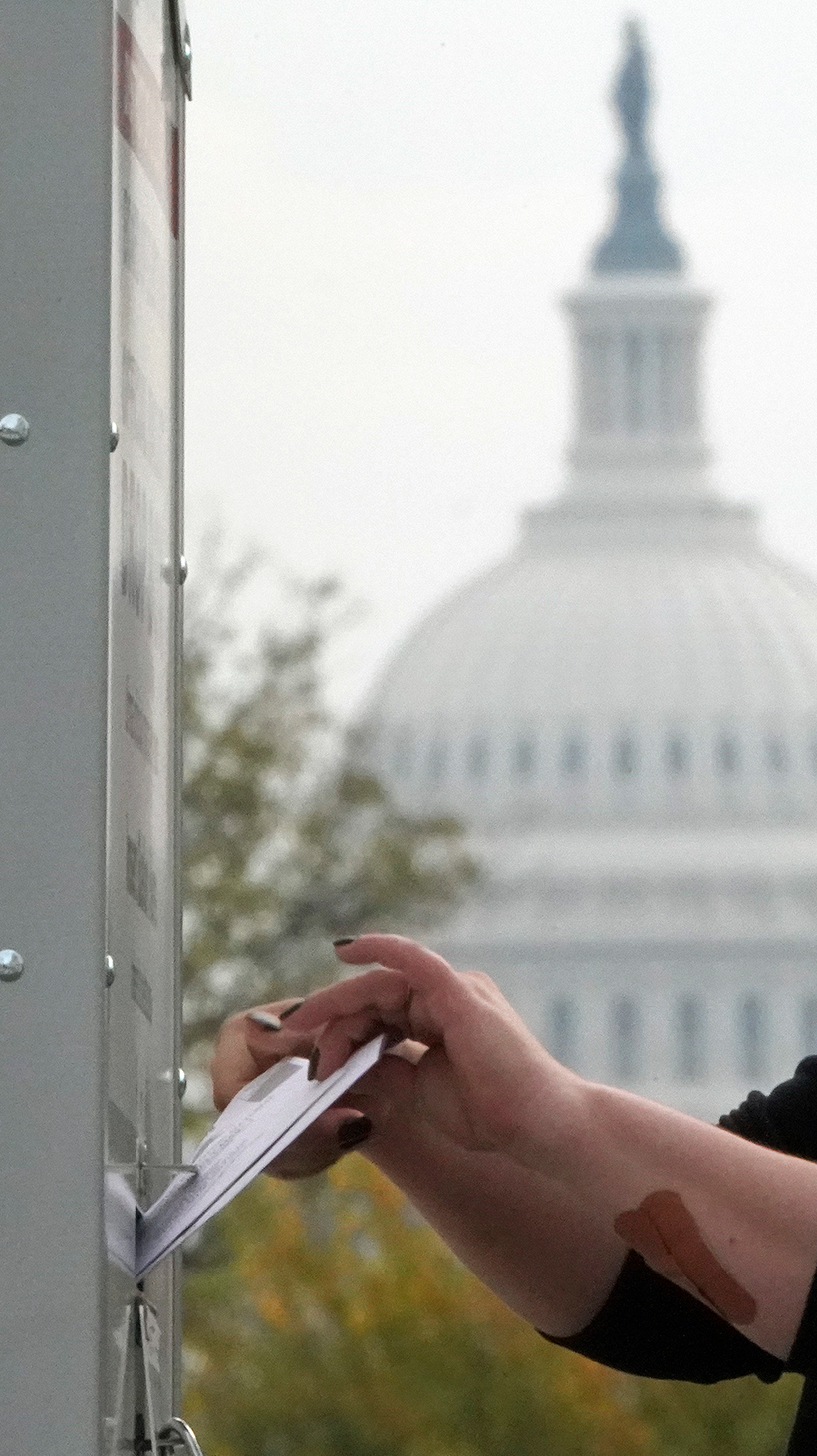 A person in a baseball cap slides a ballot into an outdoor ballot box, with the US Capitol in the background.