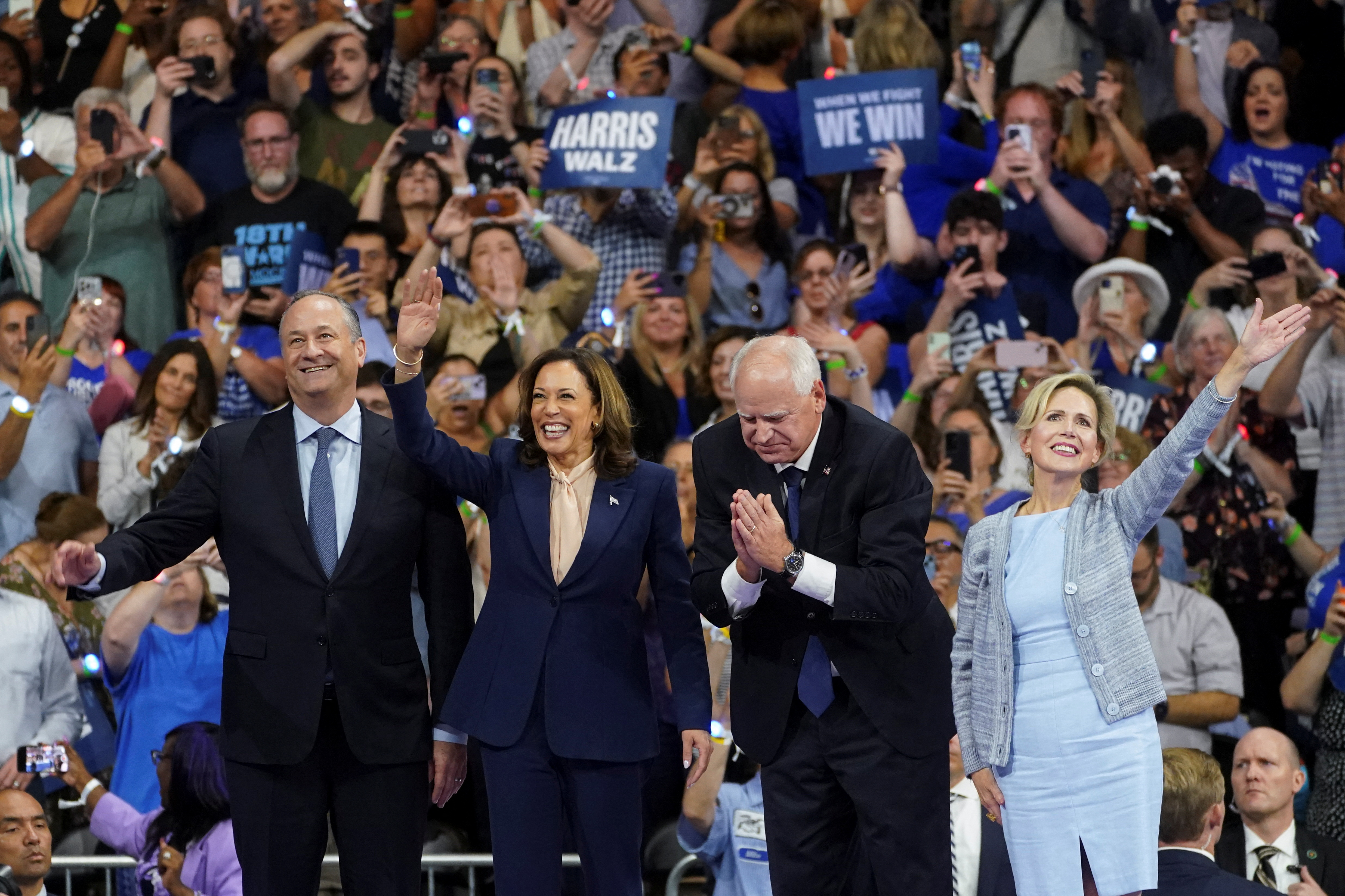 Kamala Harris and running mate Tim Walz on stage with their spouses. They are smiling and waving. There are crowds of happy people behind them.
