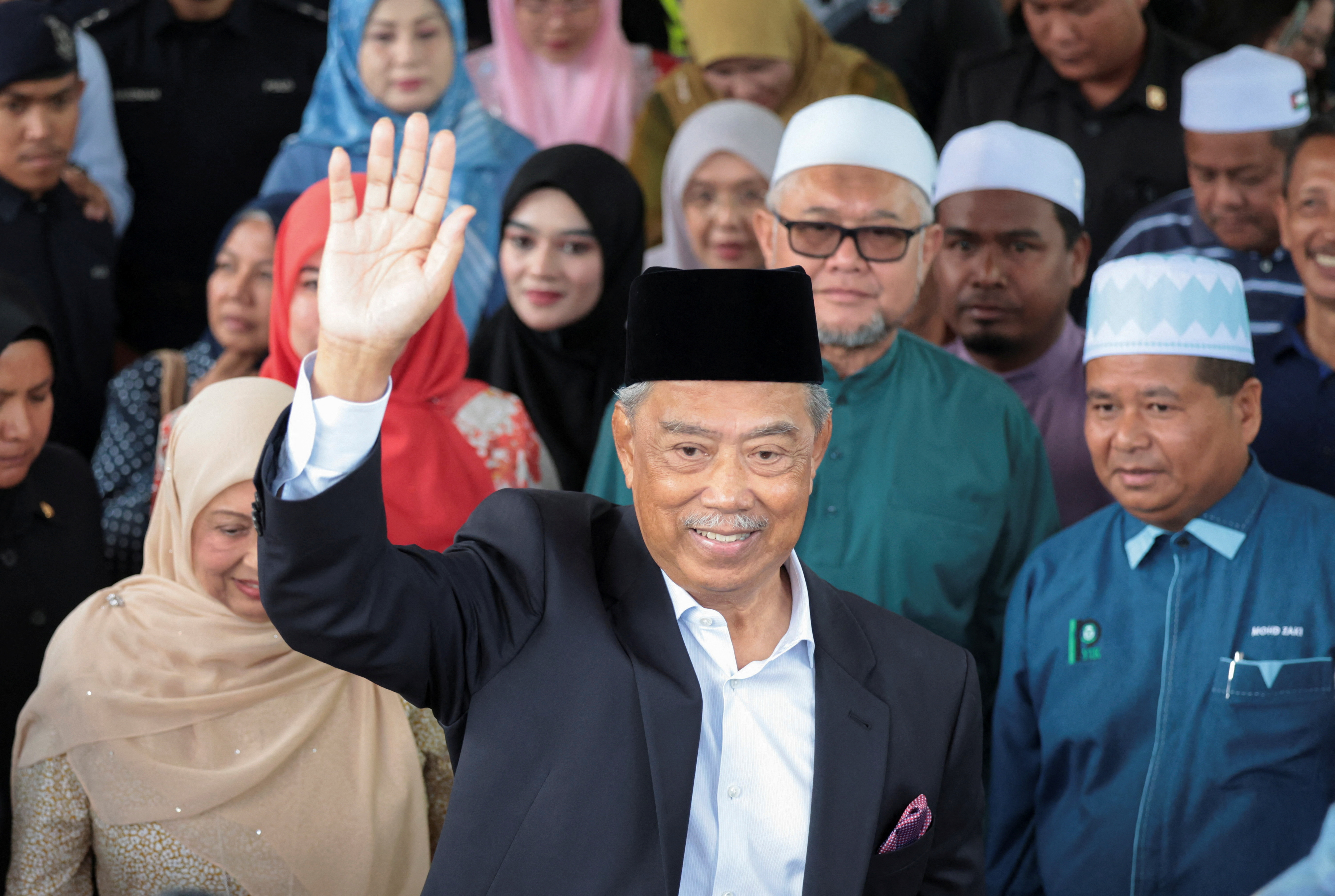 Muhyiddin leaving court. He is wearing a dark suit and a black songkok. He is waving and smiling. Supporters are behind him.