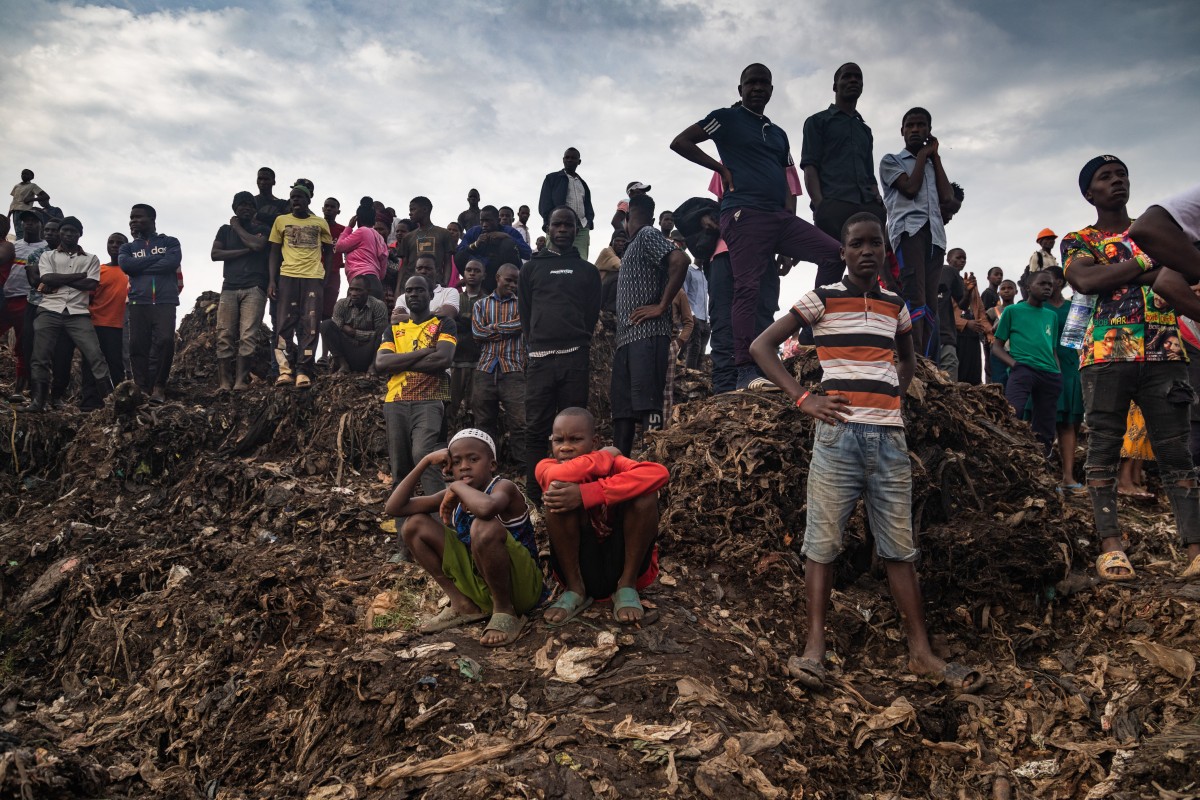 People look on as an excavator helps search for people trapped under debris after a landfill collapsed in Kampala on August 10, 2024. - Eight people including two children were killed when mountains of garbage collapsed at a landfill in the Ugandan capital Kampala on August 10, the city authority said. (Photo by BADRU KATUMBA / AFP)