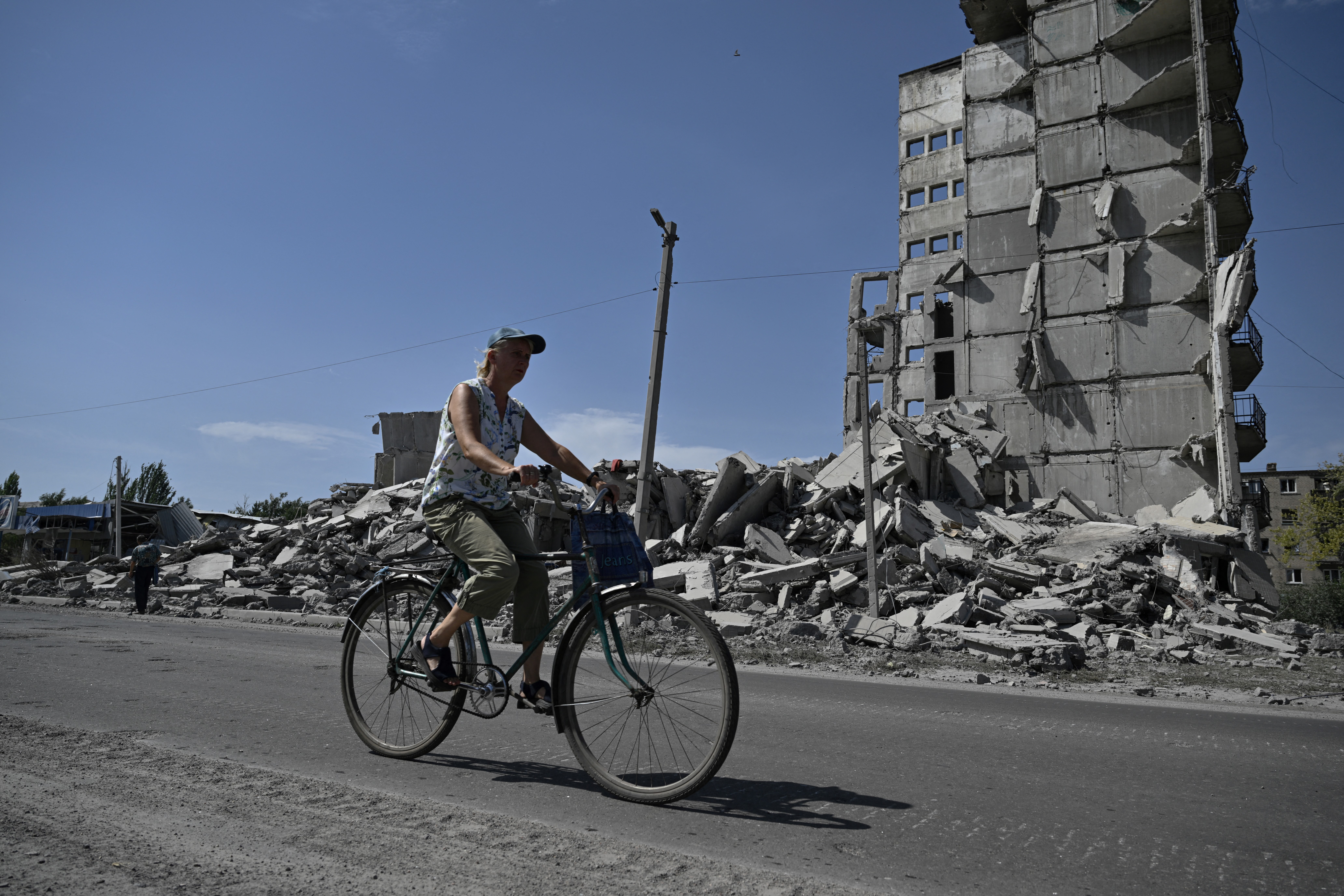 A woman rides her bicycle amid the ruins of war in Myrnohrad. An apartment block is in ruins behind her. There is rubble al around.