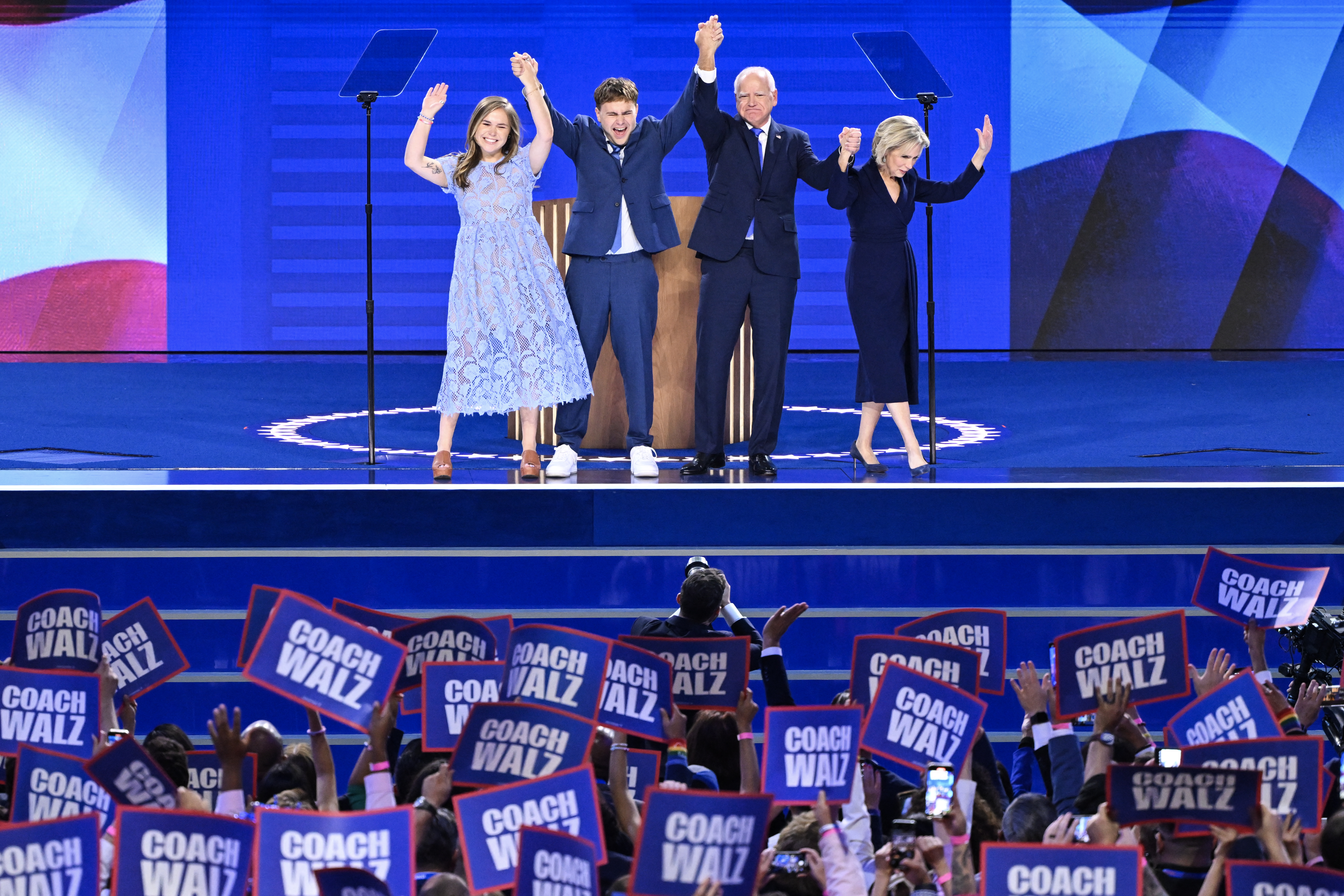 Tim Walz on stage with his wife and two children, They are smiling and waving. Poeple in front are waving Walz banners