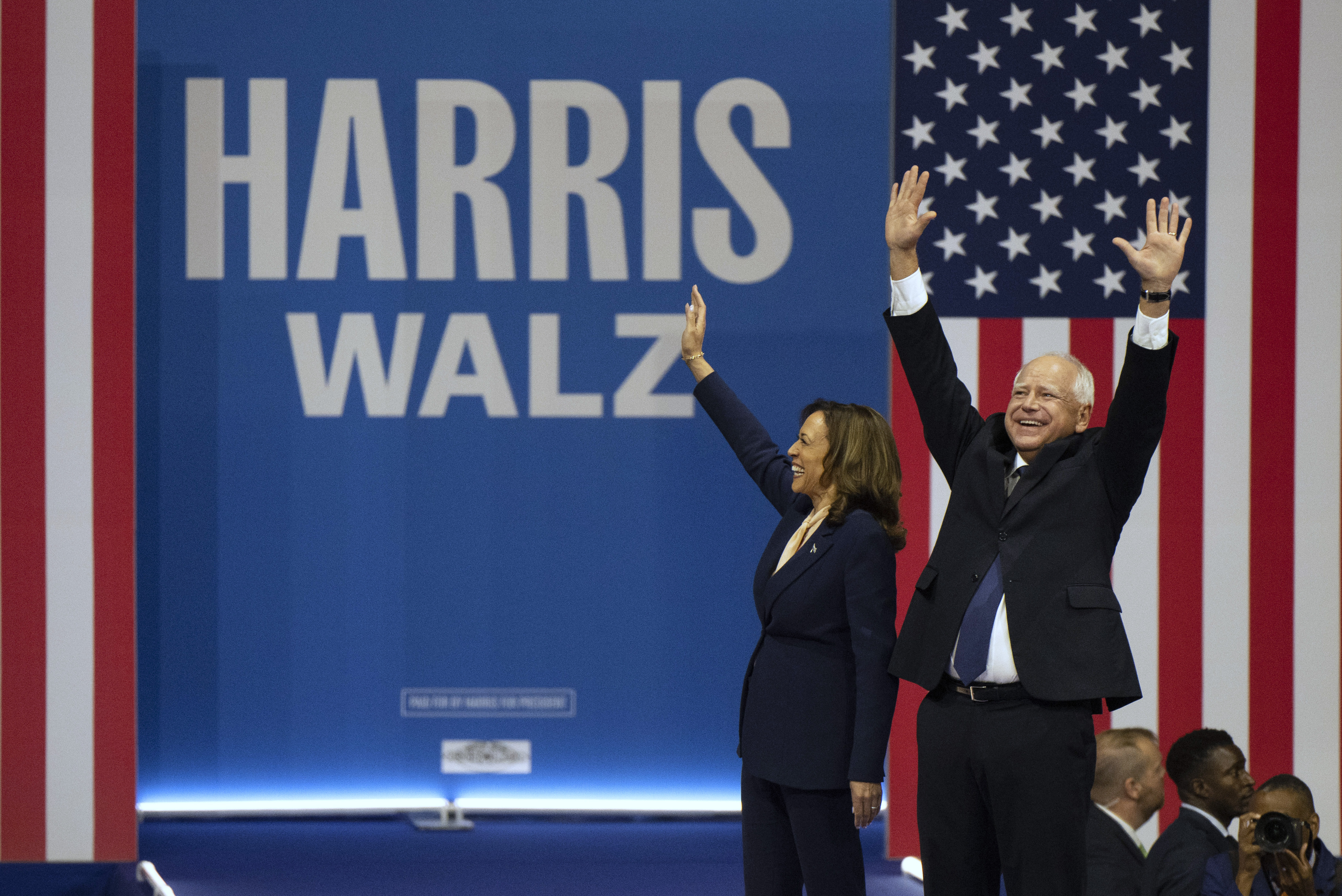 Tim Waltz and Kamala Harris raise their arms on stage in front of their names.