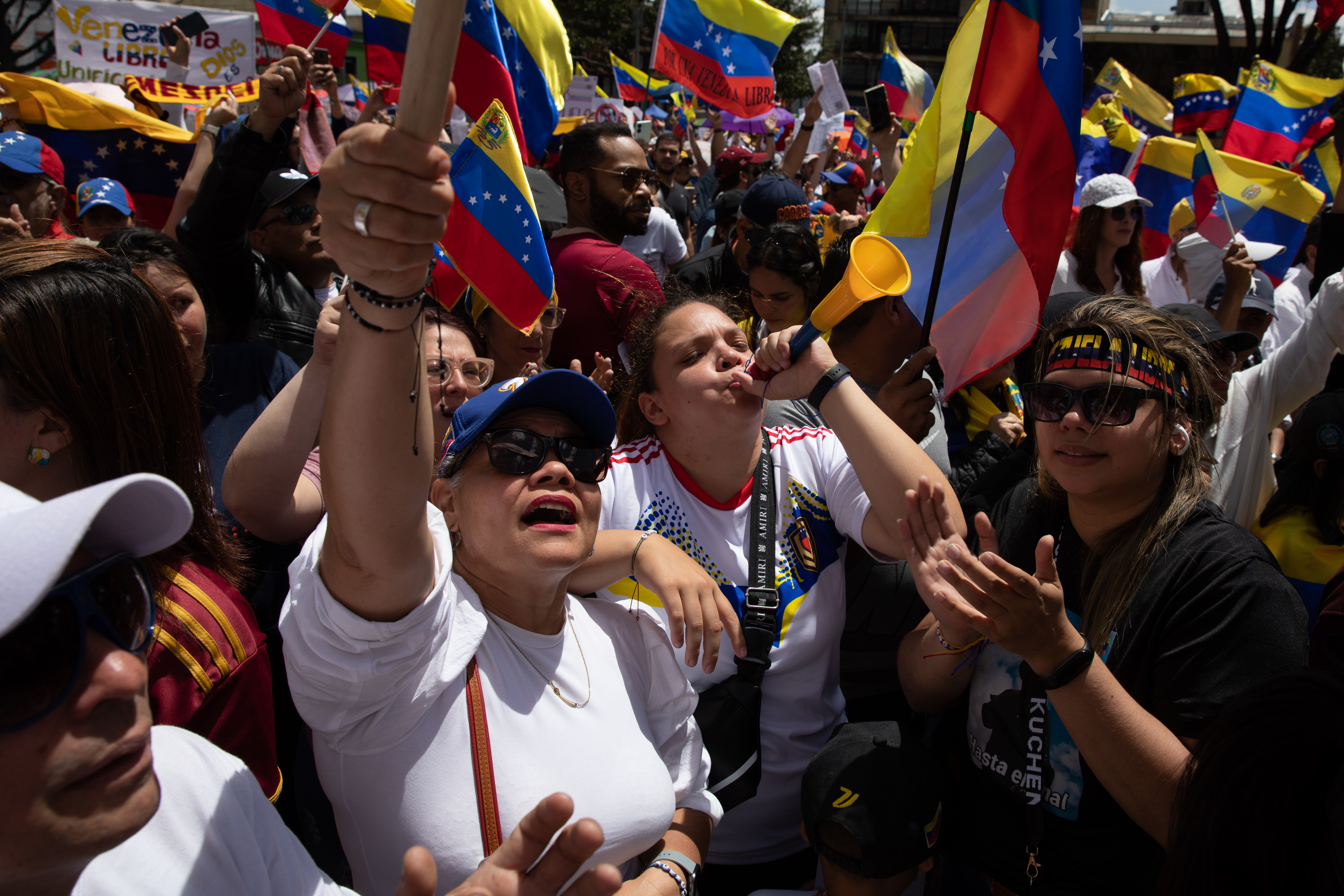 Protesters in Bogota, Colombia, crowd the street, carrying Venezuelan flags and waving their arms in the air. One woman blows a plastic horn.