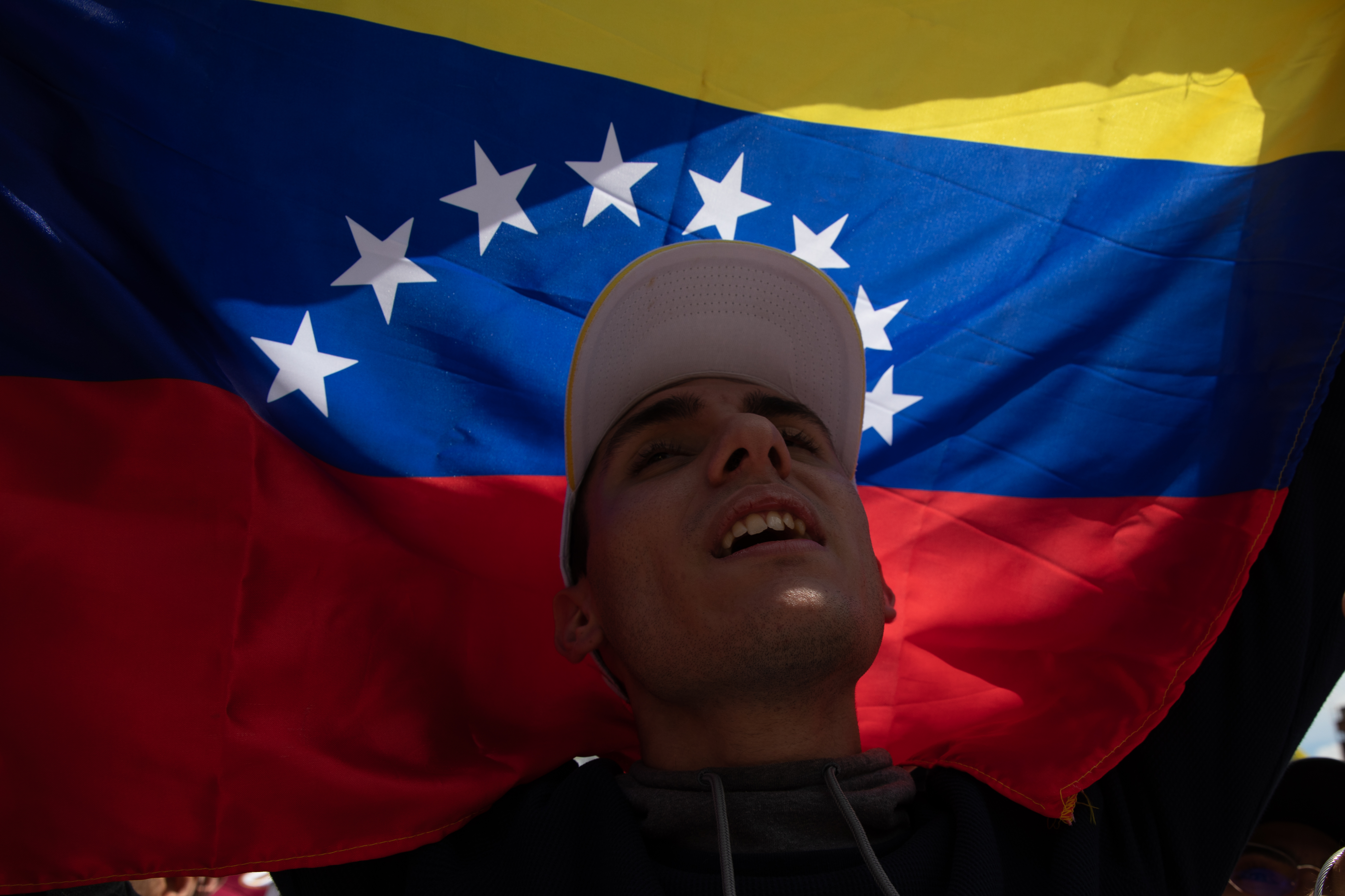 A man in a baseball cap raises a Venezuelan flag above his head, its breadth blocking out the sun behind him.