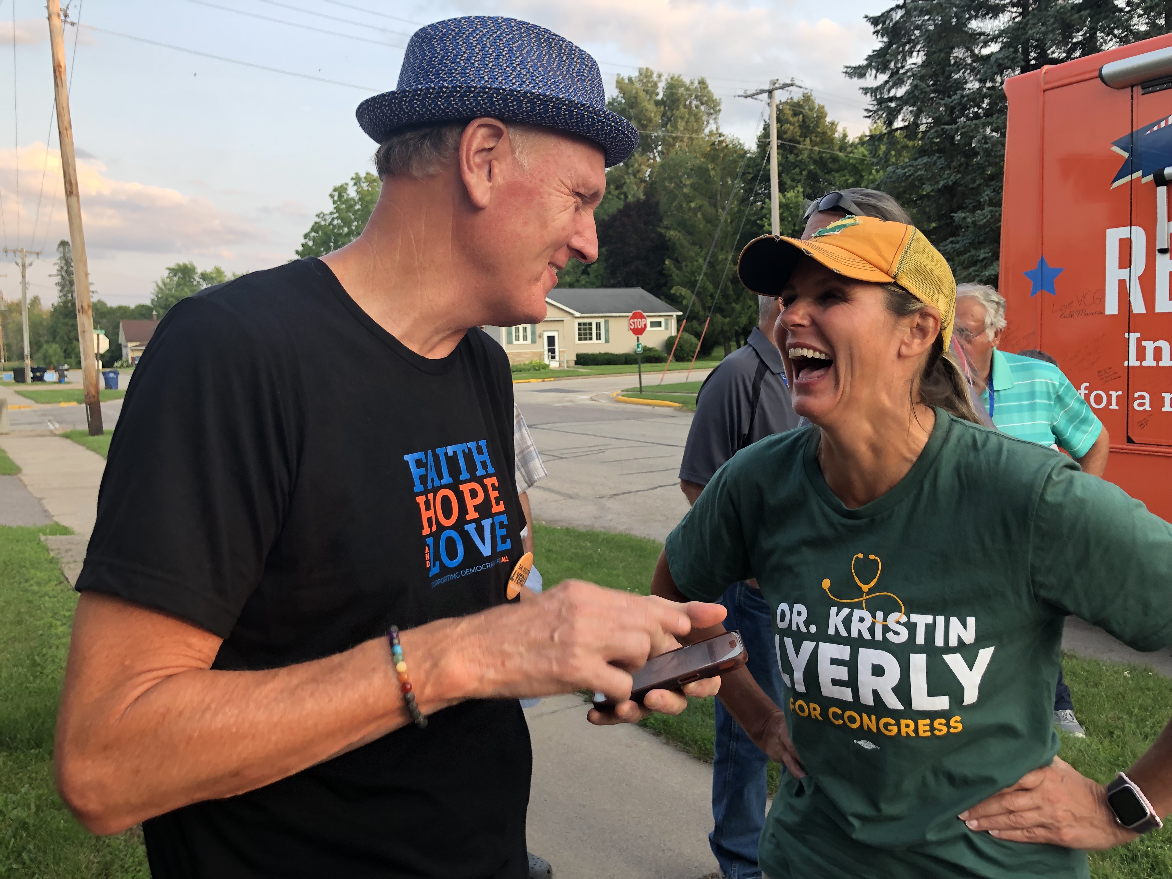 A man in a fedora talks to Kristin Lyerly with a cellphone in his hand. Lyerly — dressed in the colors of the Green Bay Packers — laughs with her hands on her hips.