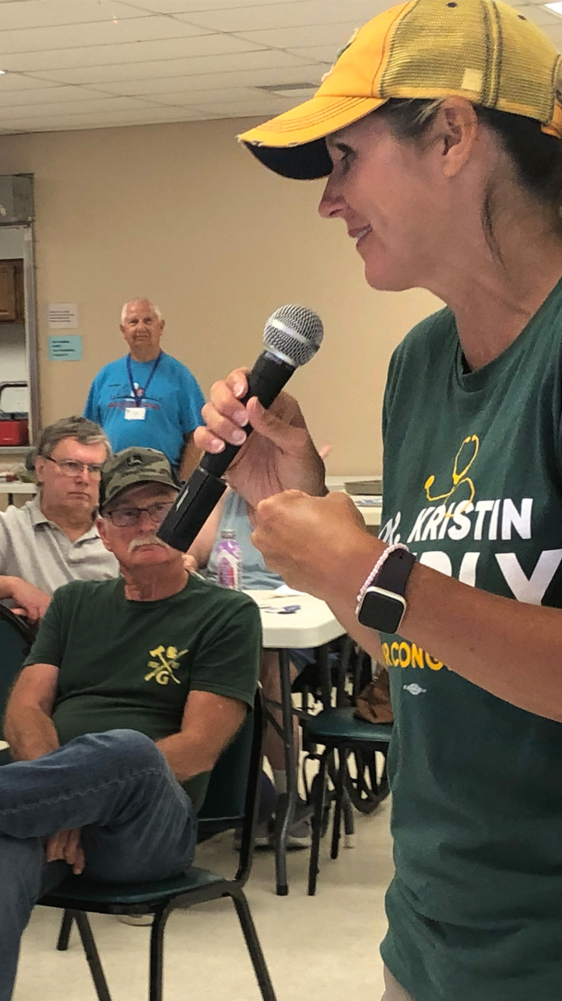 Kristin Lyerly, dressed in a yellow baseball cap and green shirt, speaks into a microphone in front of a room of voters.