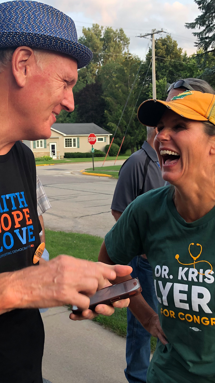 A man in a fedora talks to Kristin Lyerly with a cellphone in his hand. Lyerly — dressed in the colors of the Green Bay Packers — laughs with her hands on her hips.