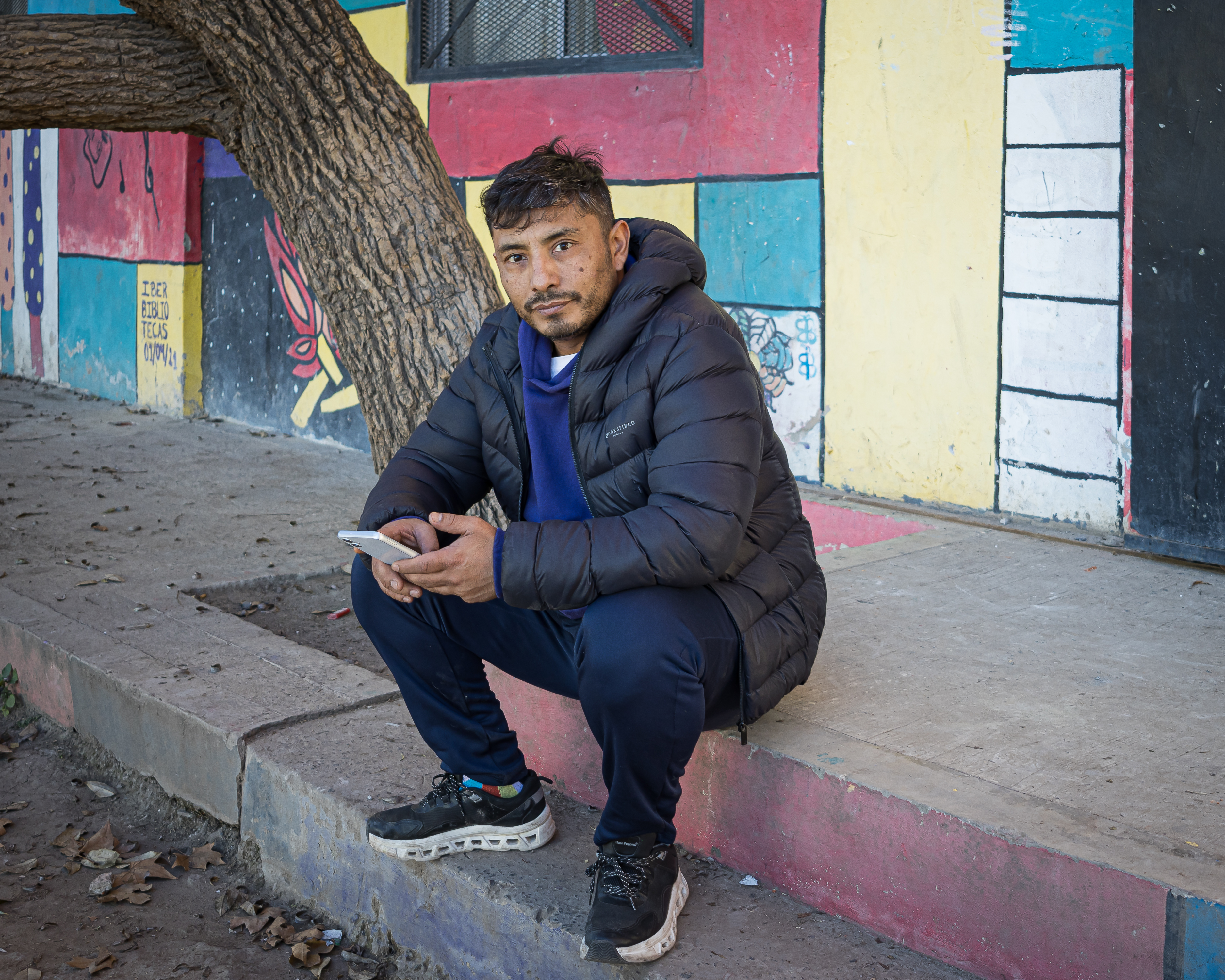 Waldemar Cubilla sits on a street curb, dressed in a quilted, hooded jacket. In his hands is a cellphone.