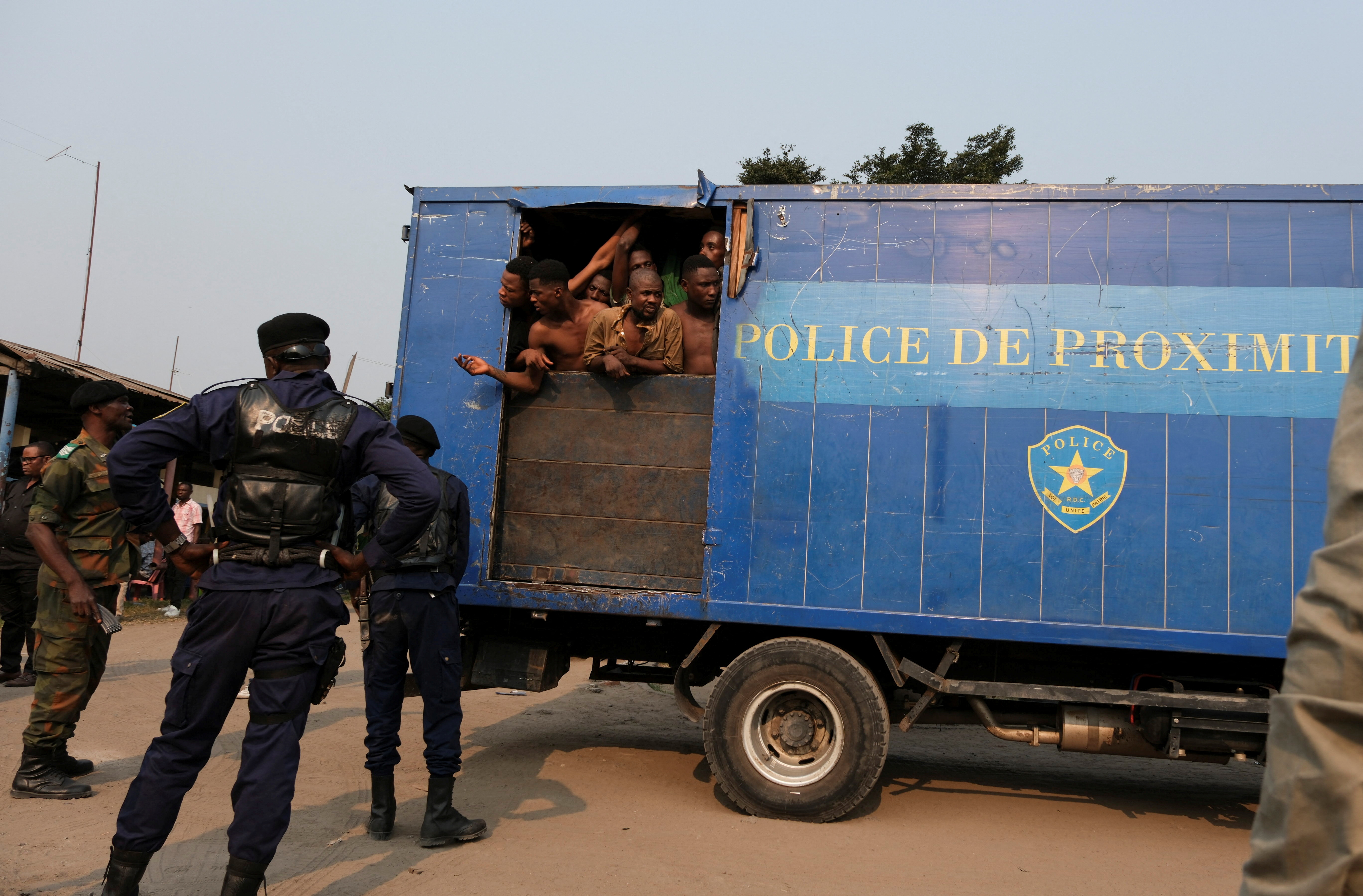 Prisoners arriving at Makala prison in Kinshasa. They are packed into a blue truck. Some are looking out of an opening at the back. Police are on the street
