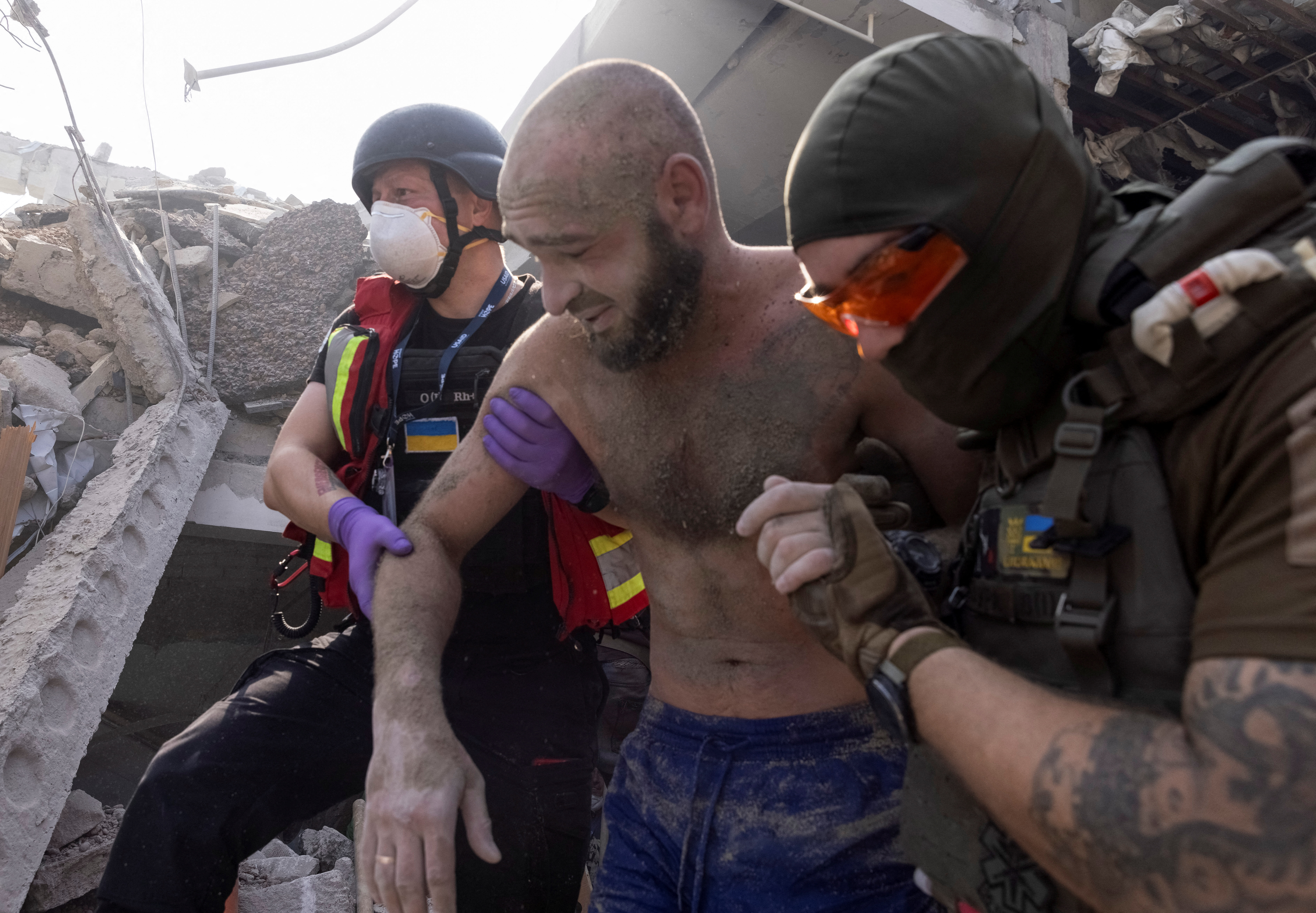 A man being escorted from the rubble of a building in Kharkiv. He is covered in dust and is wearing only shorts. There are two rescuers on either side of him. There is lots of rubble behind them.