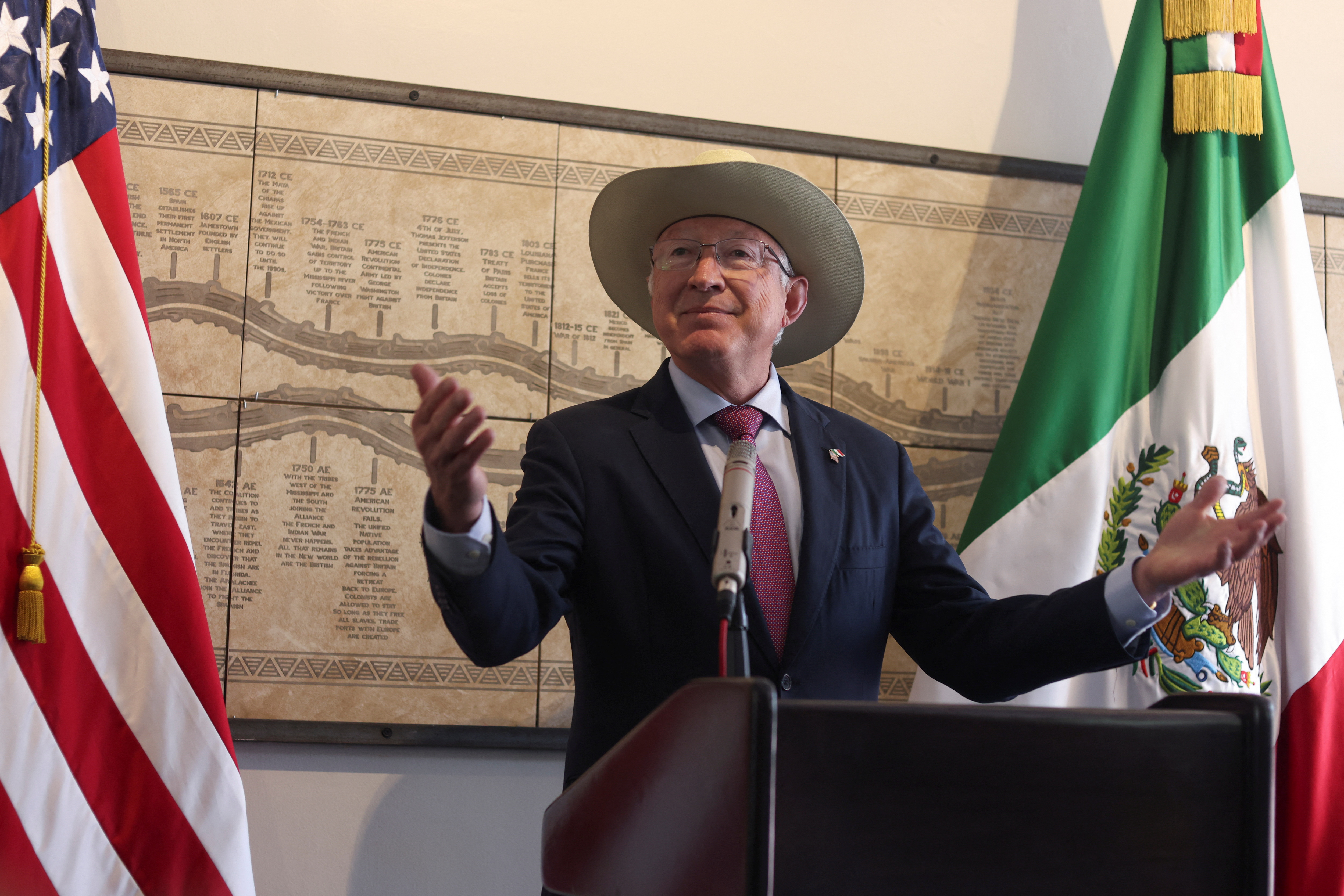 Ken Salazar, in a wide-brimmed hat behind a podium, surrounding by US and Mexican flags.