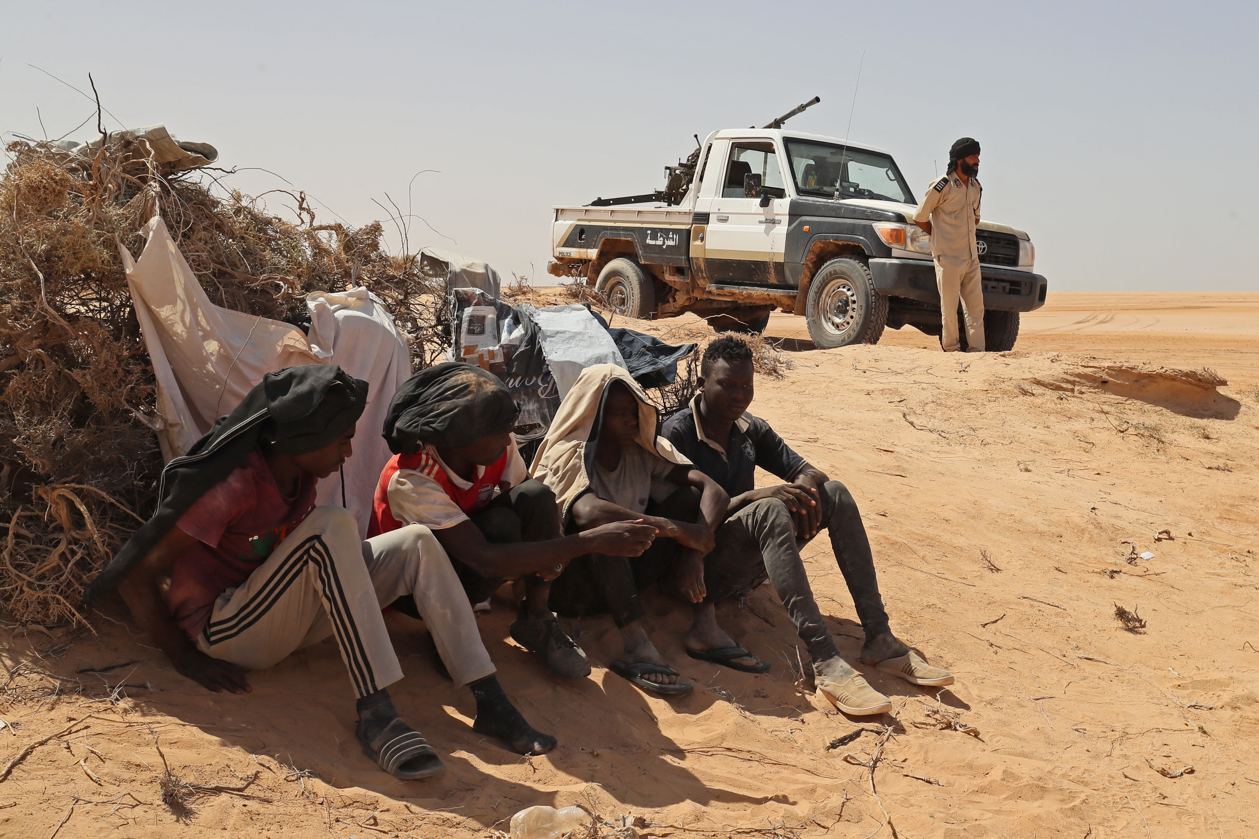 A Libyan border guard stands near migrants from sub-Saharan African countries who claim to have been abandoned in the desert by Tunisian authorities without water or shelter, during a rescue operation in an uninhabited area near the border town of Al-Assah on July 16, 2023. Hundreds of migrants from sub-Saharan African countries were forcibly taken to desert and hostile areas bordering Libya and Algeria after unrest in early July in Sfax, Tunisia's second-largest city. (Photo by Mahmud Turkia / AFP)