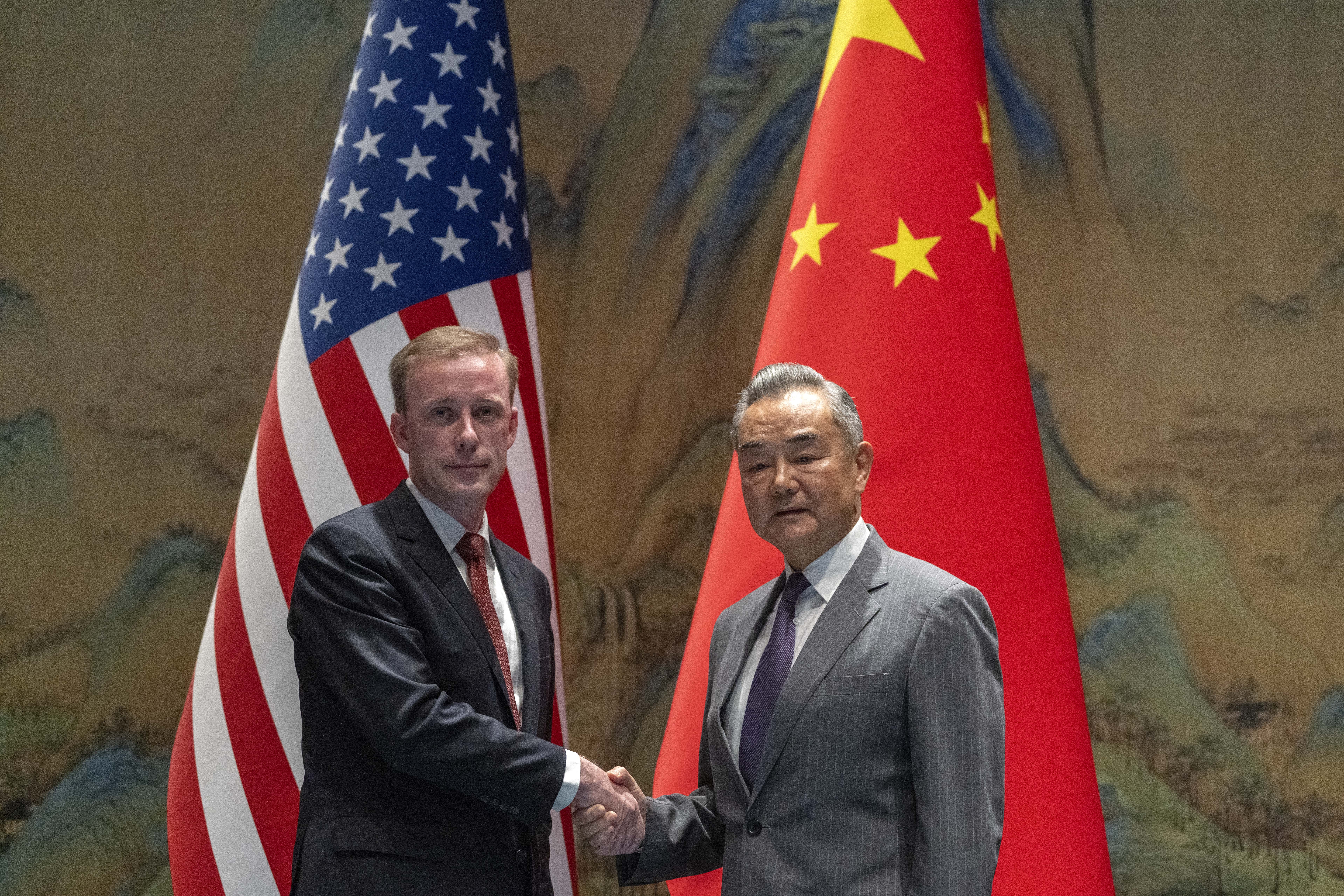 Jake Sullivan and Wang Yi shaking hands. They are standing in front of the flags of their respective countries.