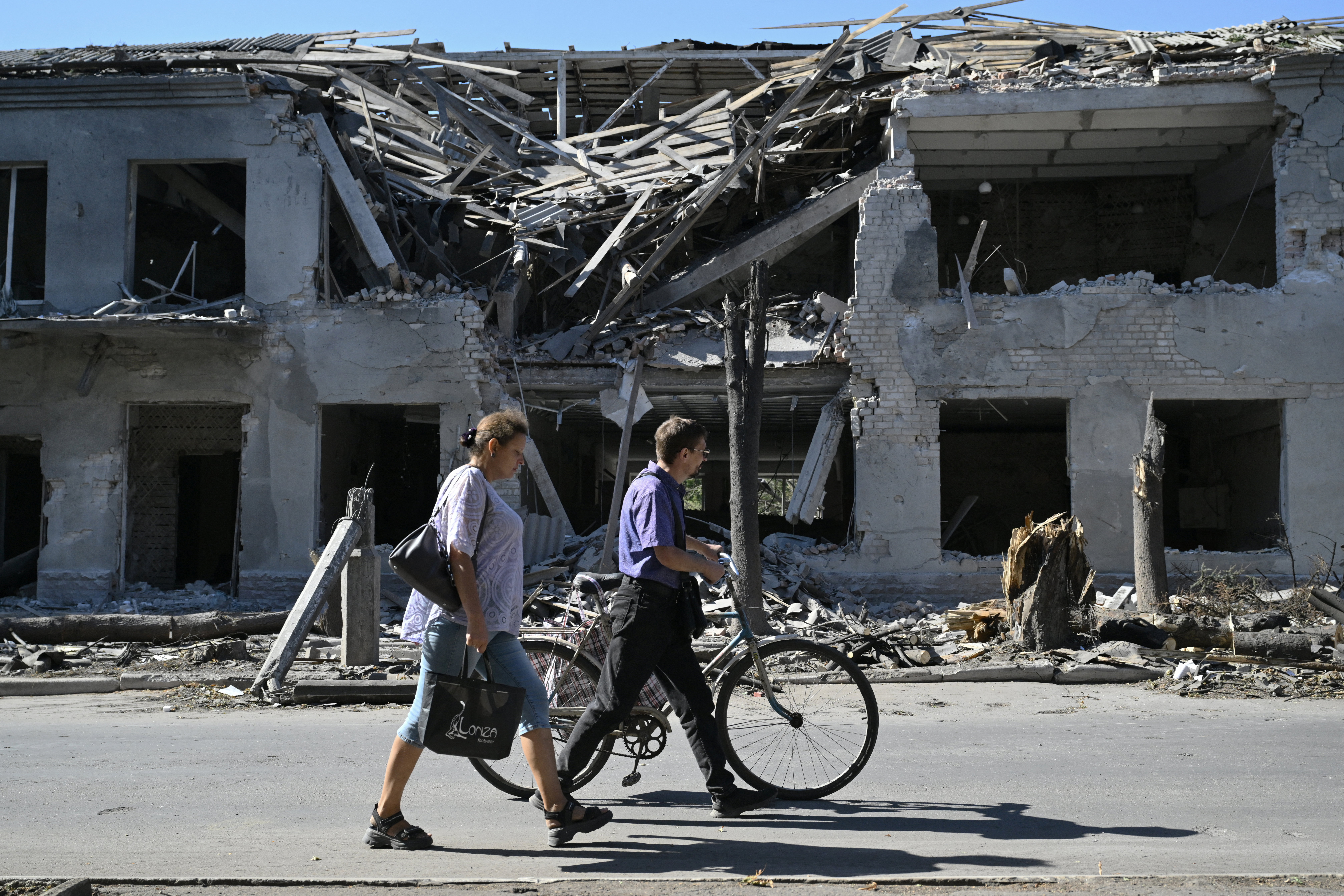 Two people walking past a badly damaged building in Pokrovsk. One of the people is pushing a bicycle.