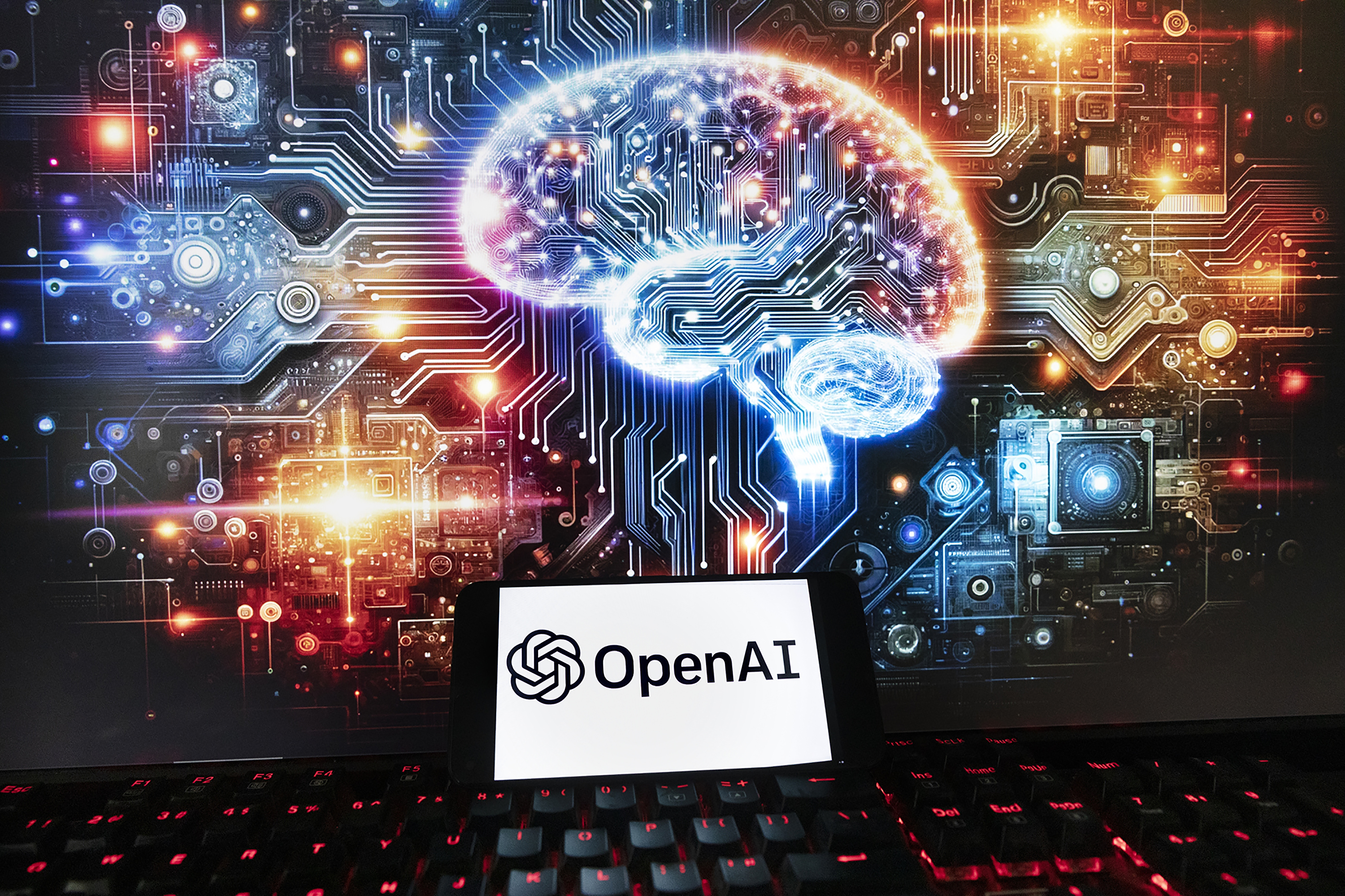 The OpenAI logo is displayed on a cell phone with an image on a computer monitor generated by ChatGPT's Dall-E text-to-image model