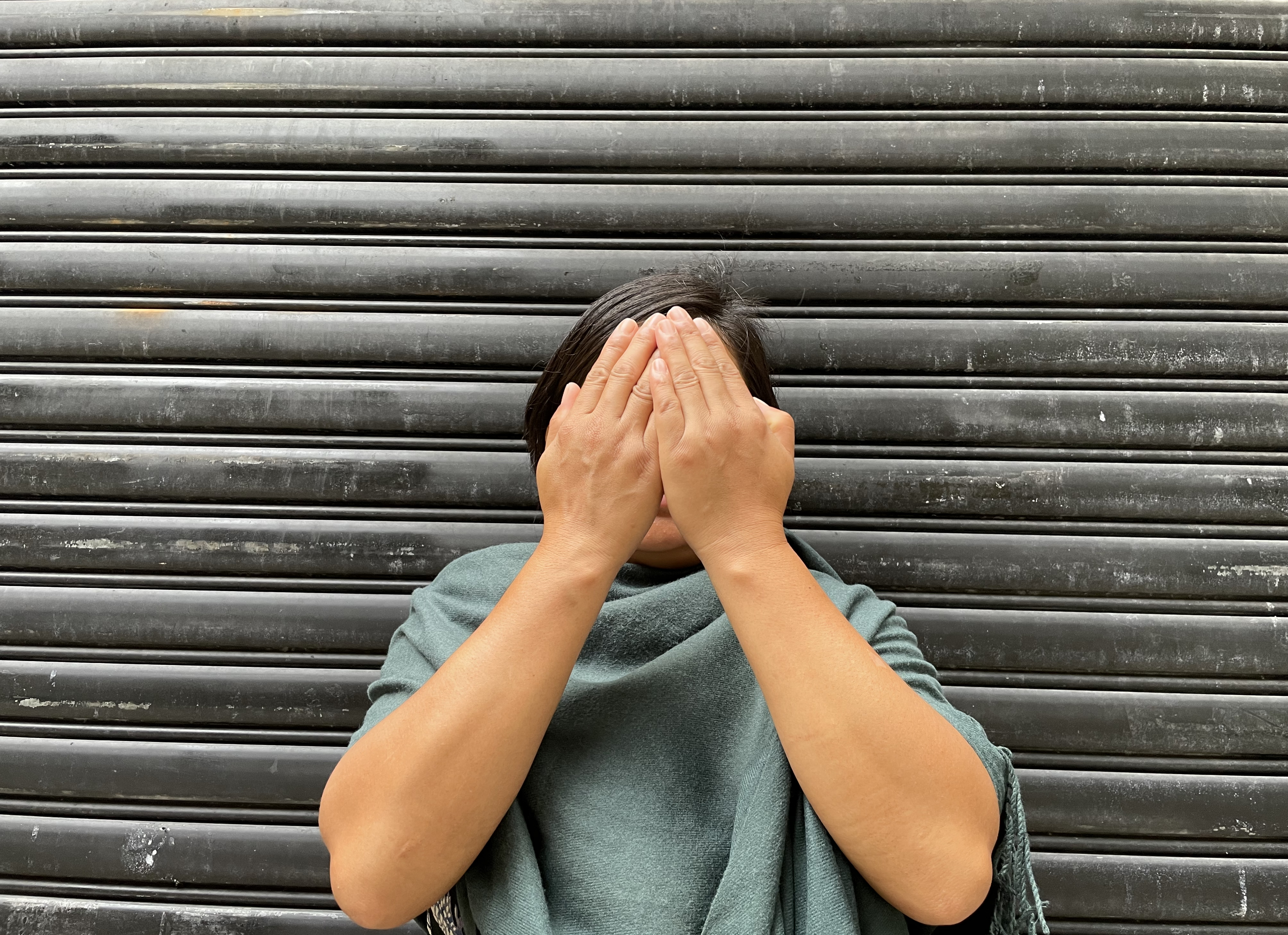 X with her hands over her face to protect her identity. She is standing in front of a metal shutter