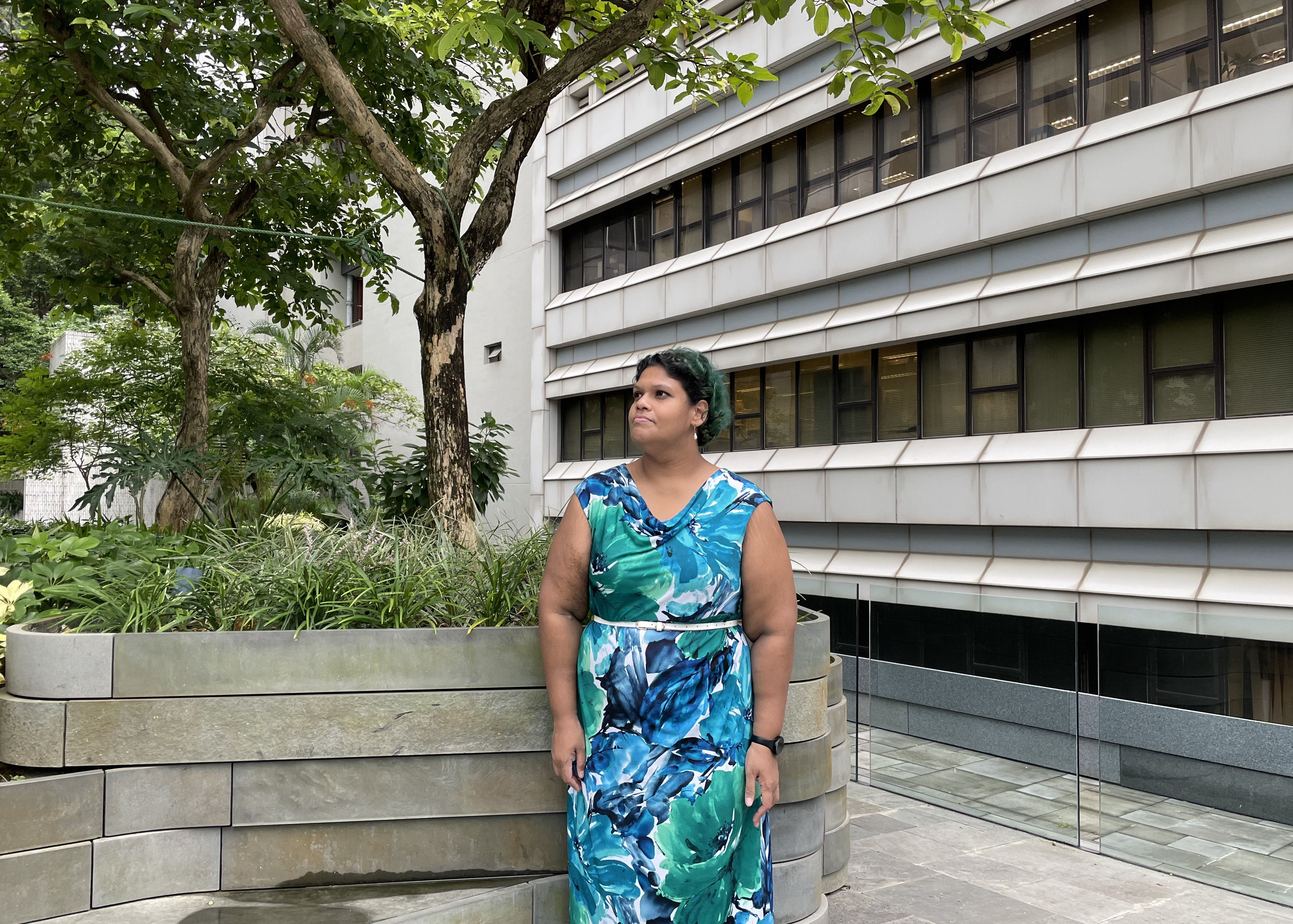 Manisha Wijesinghe, She is in a aquamarine dress standing outside in a courtyard. She has short blue hair.