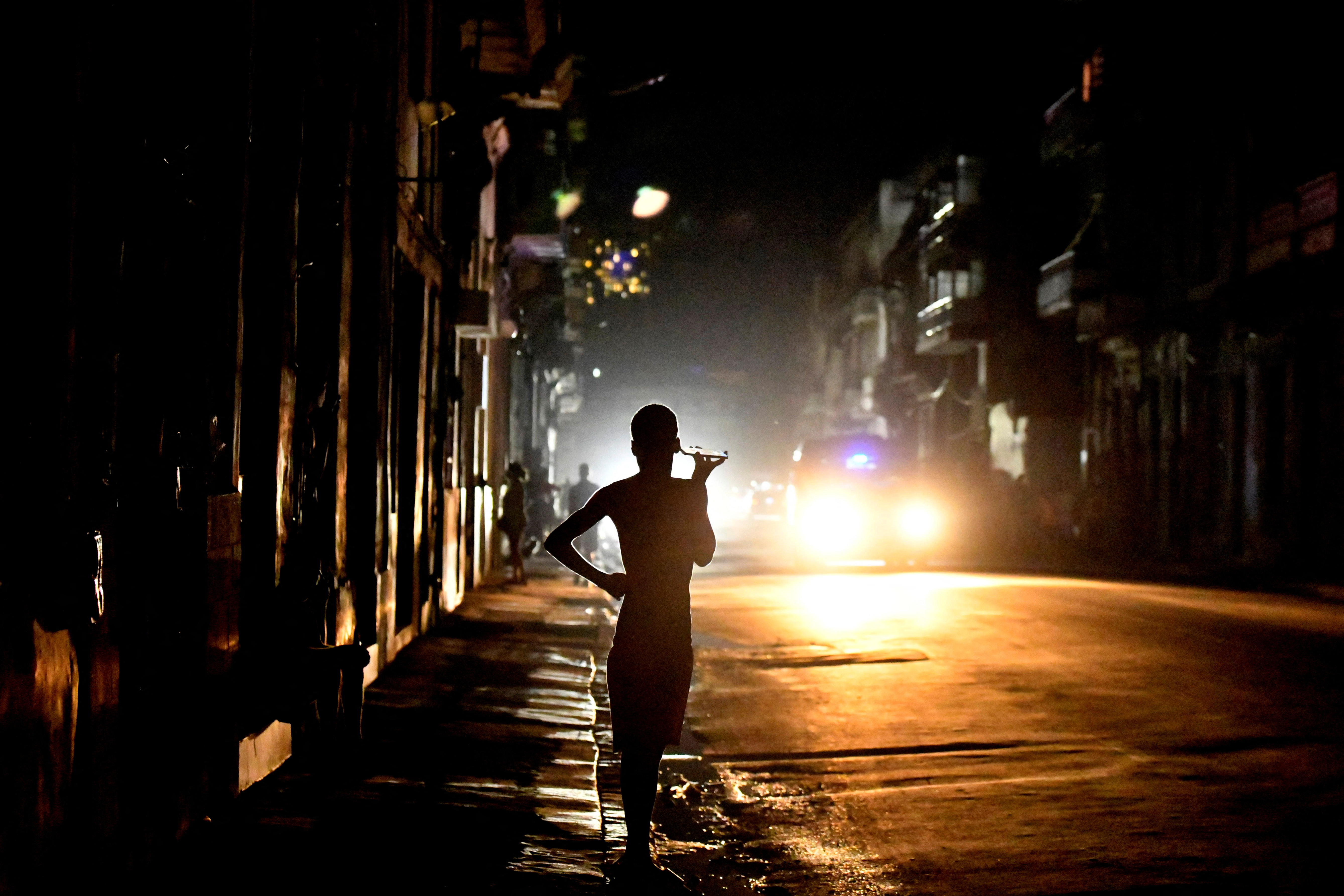 A darkened silhouette holds a cellphone up to her ear during a blackout. A car light can be seen on the darkened street.