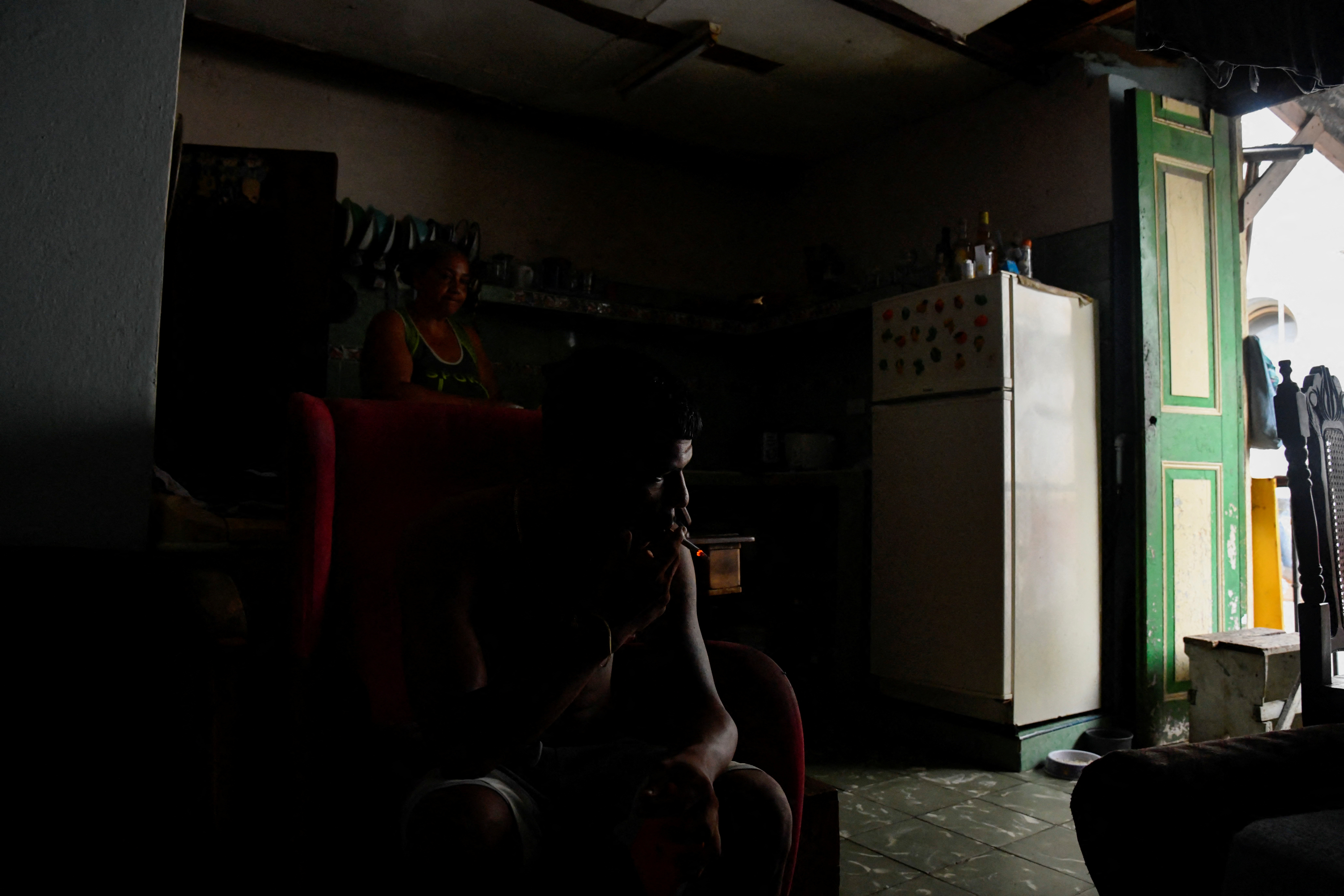 A person smokes a cigarette in his darkened home during a blackout, while sun pours in through an open door.