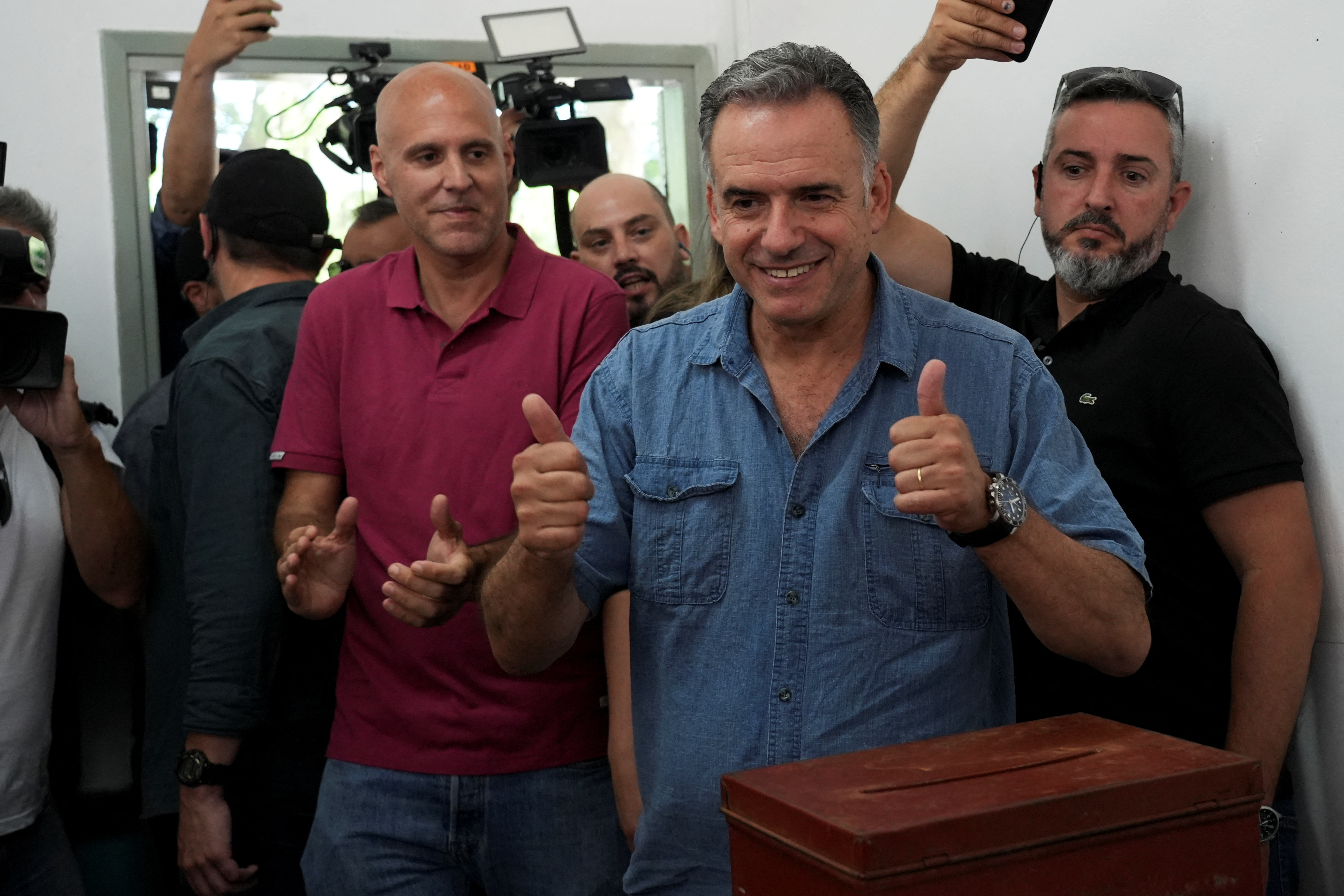 Yamandu Orsi gives the media two thumbs up after casting his ballot. Photographers crowd behind him with cameras in the air.