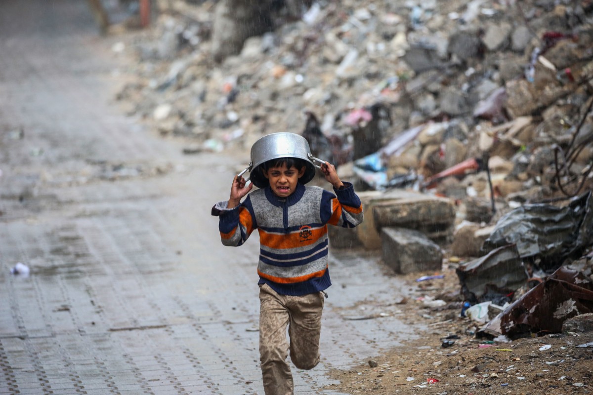 A displaced boy covers his head with a pan as he runs from the rain past building rubble at the Bureij refugee camp
