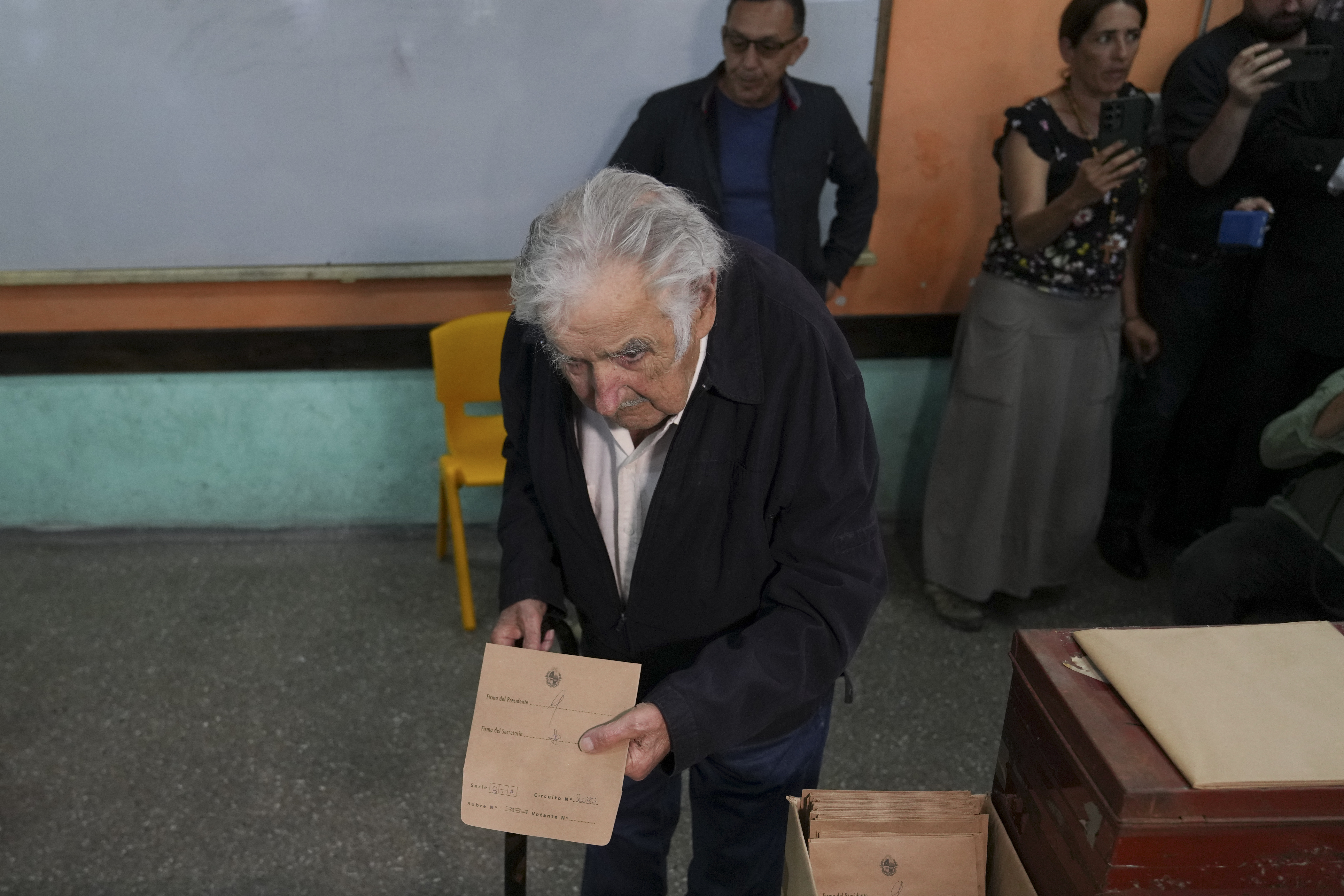 An elderly Jose Mujica is bent over, wearing a black suit, is holding the brown envelope with his vote, ready to cast. People behind him stand against a wall.