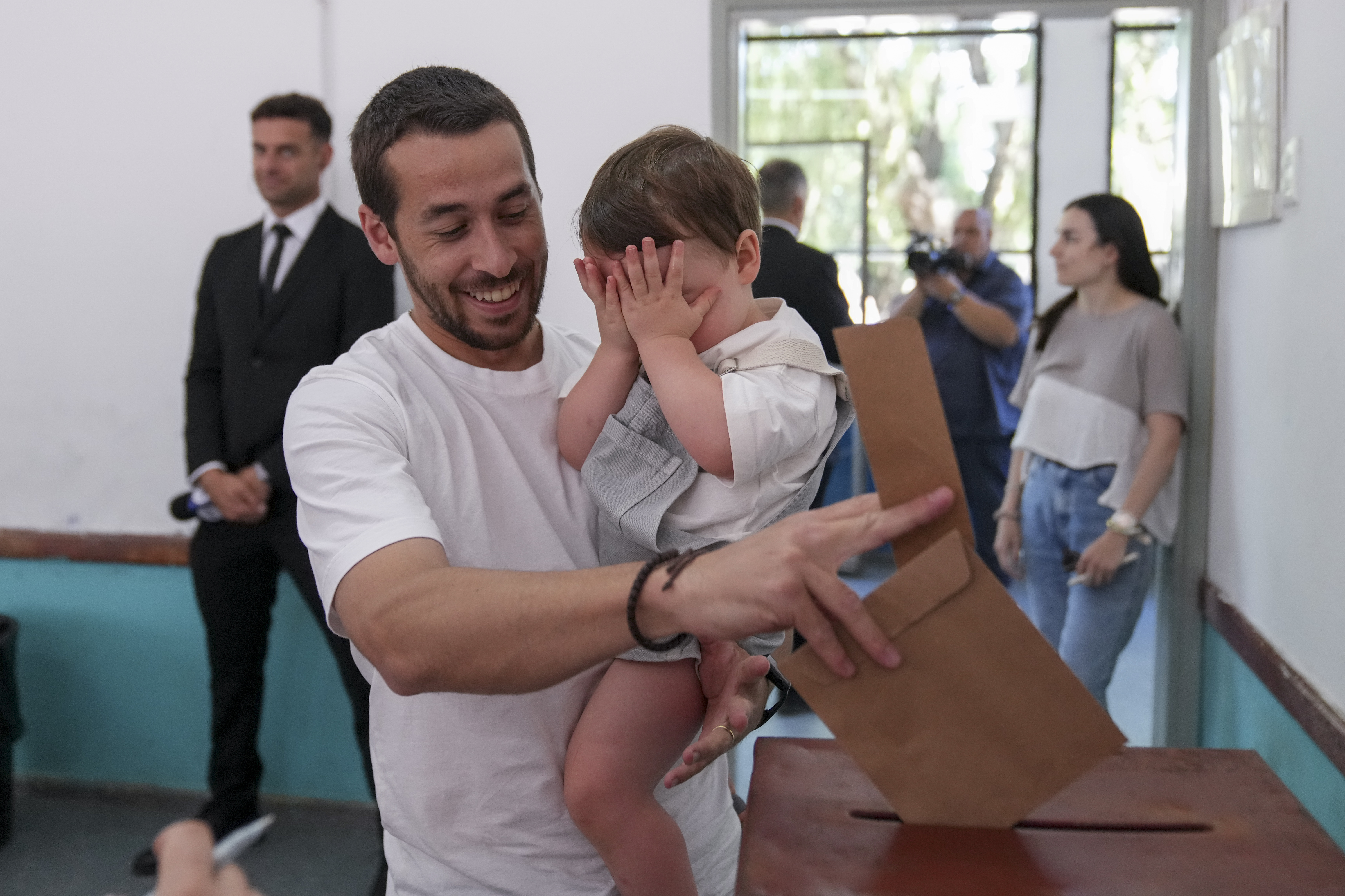 A man casts his ballot at a polling station in Uruguay, with a baby in his arms. The man is smiling while the little boy covers his face with his hands.