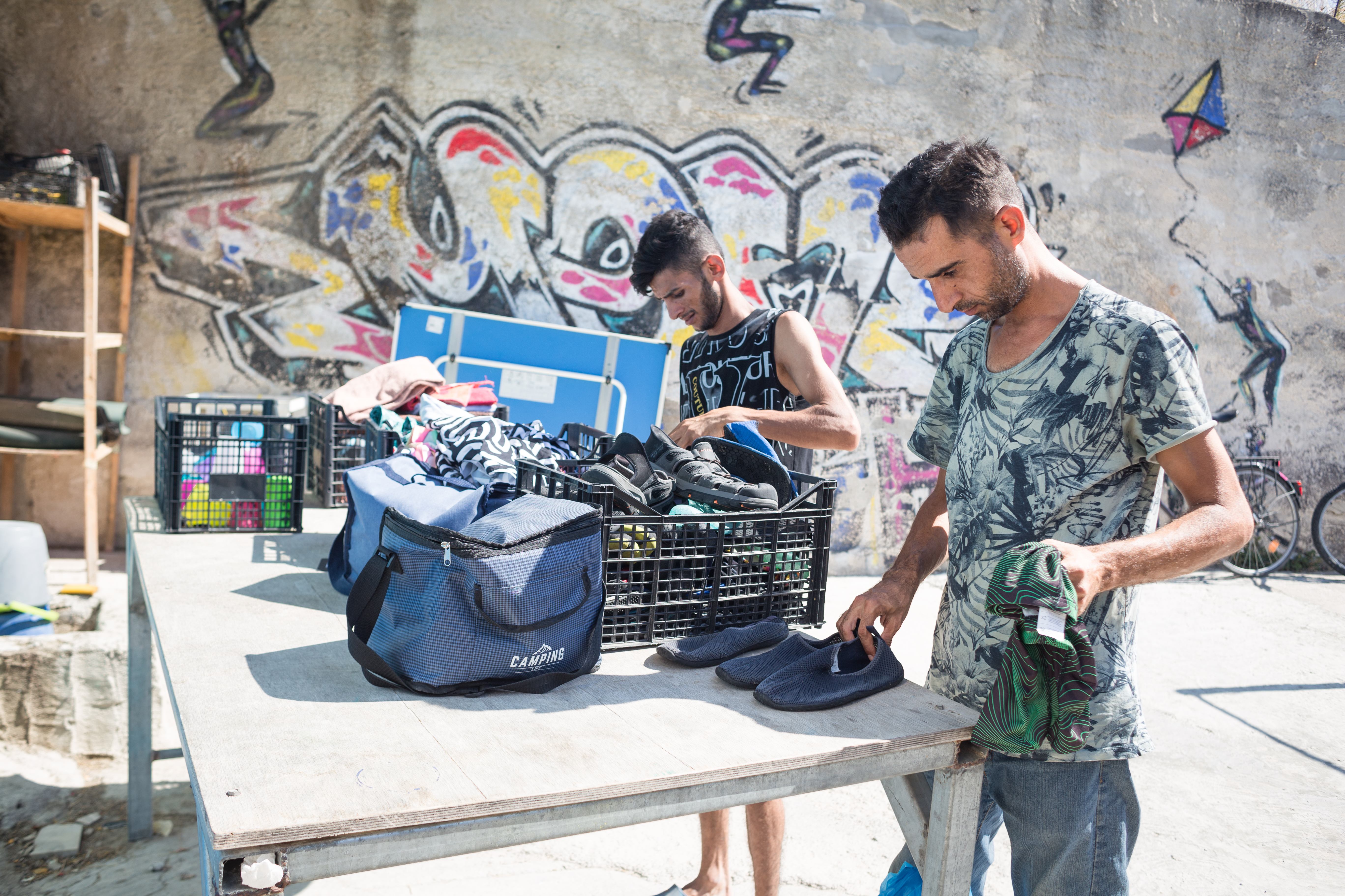 Two men look through a basket full of clothes and shoes. There is a graffitied wall behind them.