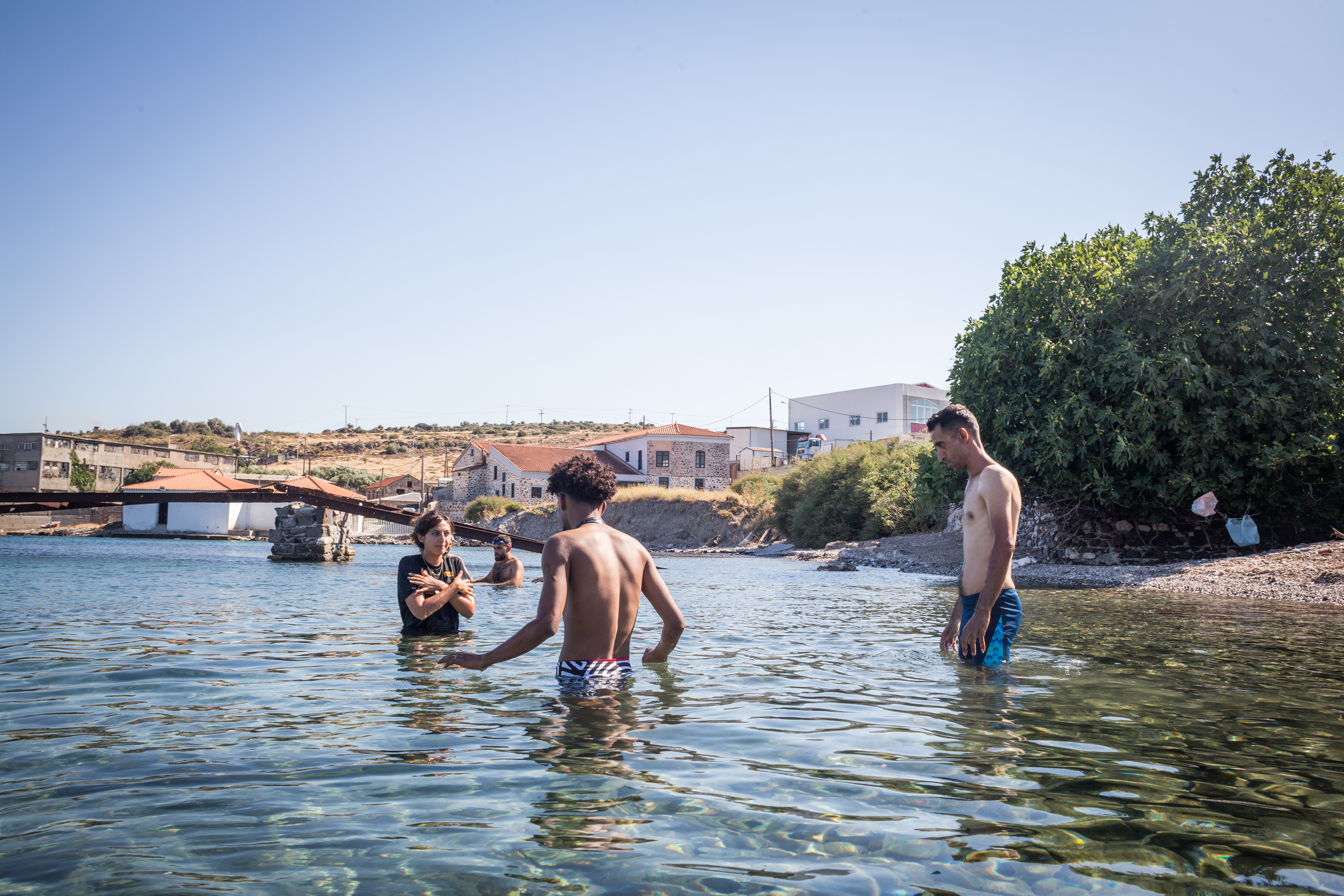 Men stand waist-deep in the water. There are houses on the shore behind them and what looks like a collapsed rail bridge going across the water.