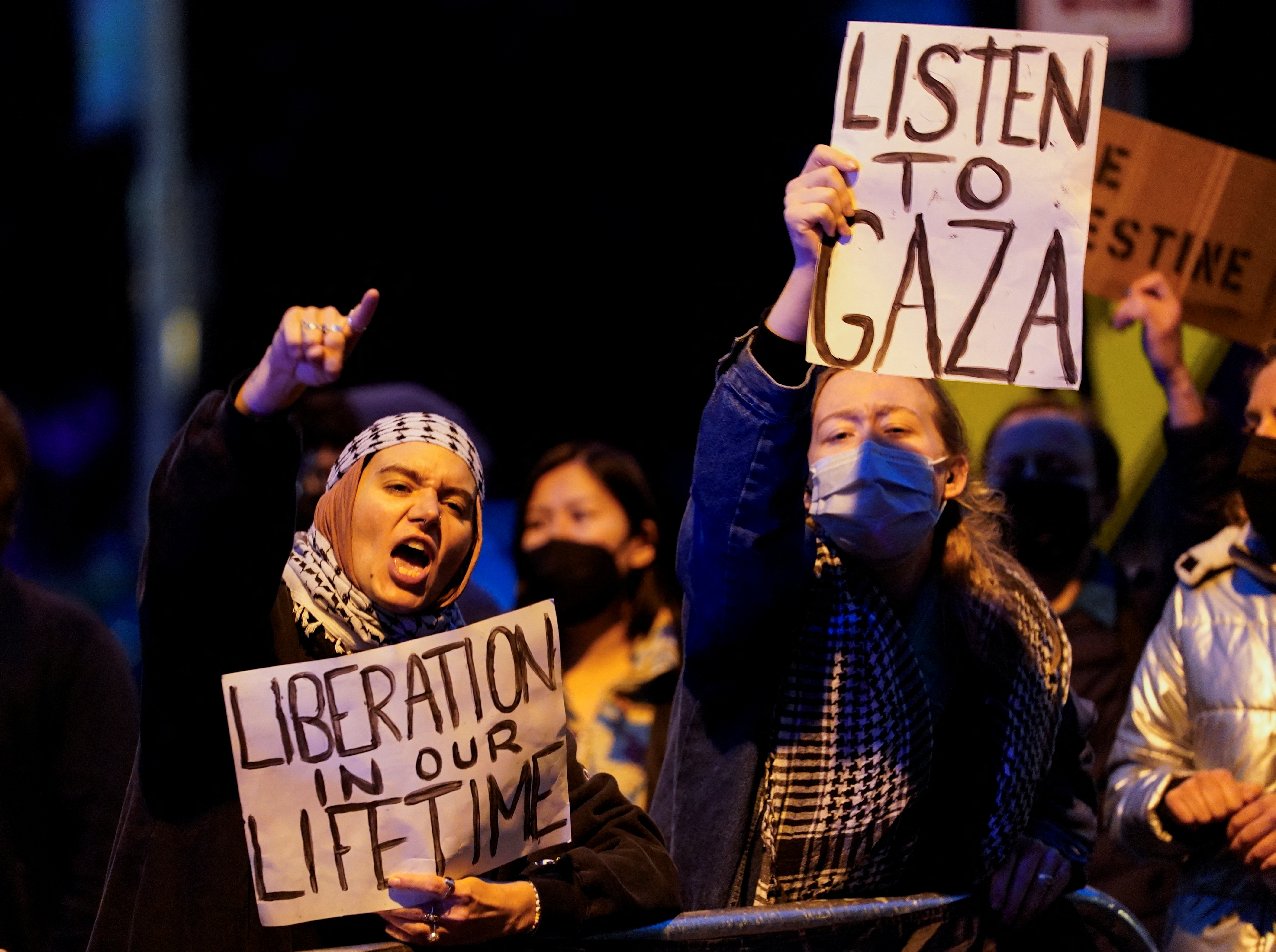 Protesters at a nighttime demonstration raise fists and hold up signs that read "Liberation in our Lifetime" and "Listen to Gaza"