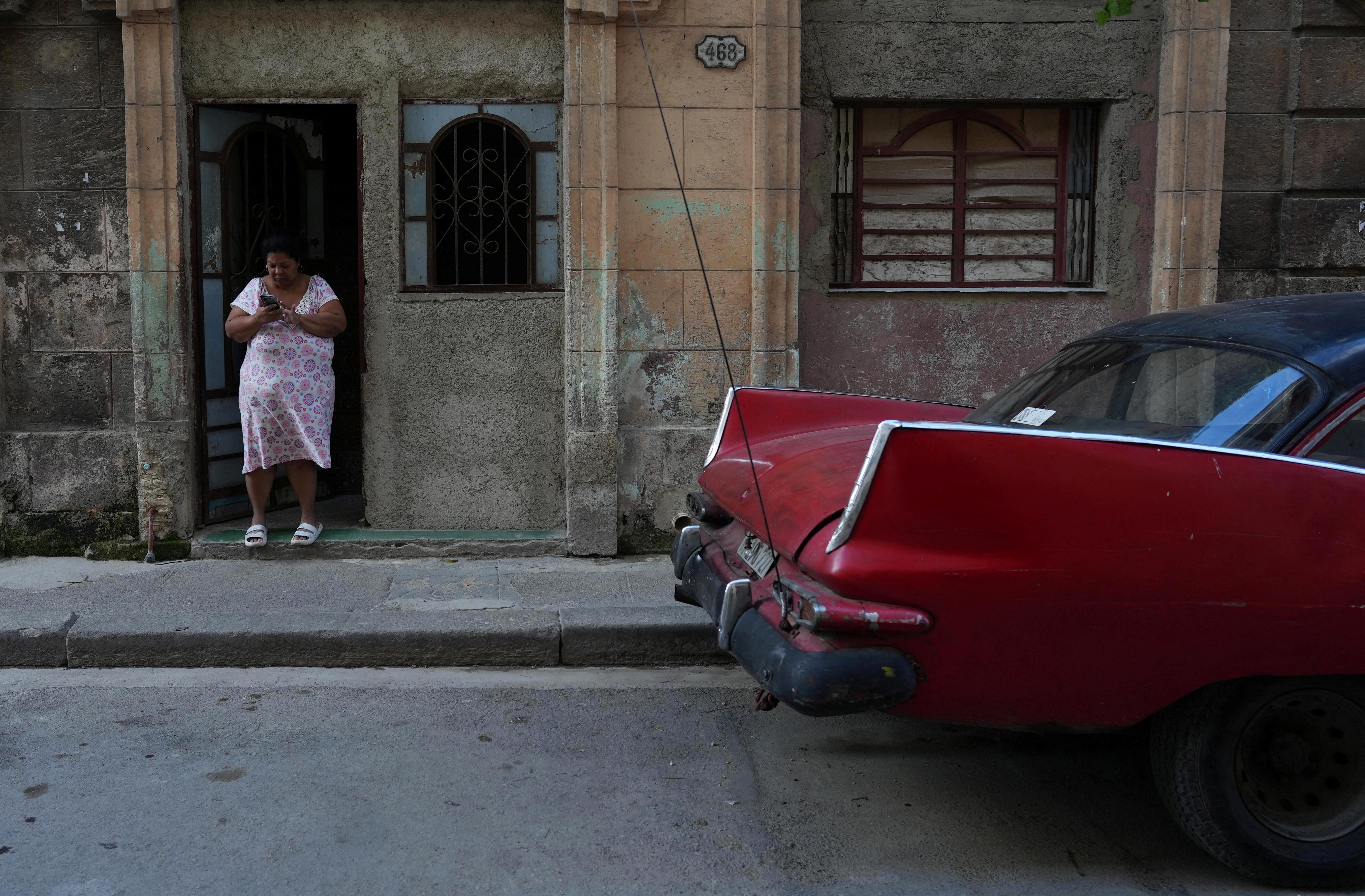 A woman checks her phone in a doorway in Havana, Cuba, as a car drives by