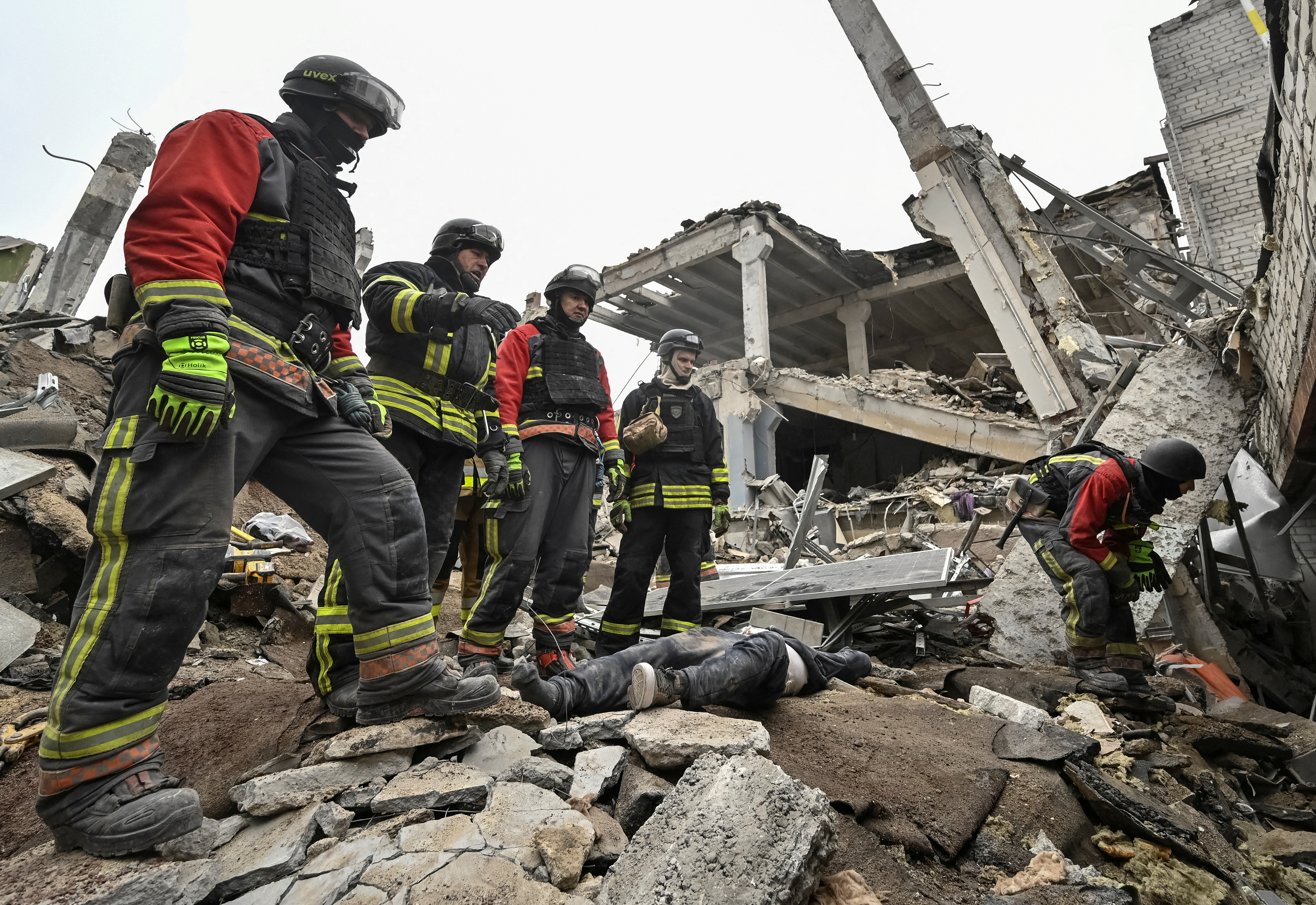 people in firefighting uniforms stand next to a body lying among rubble