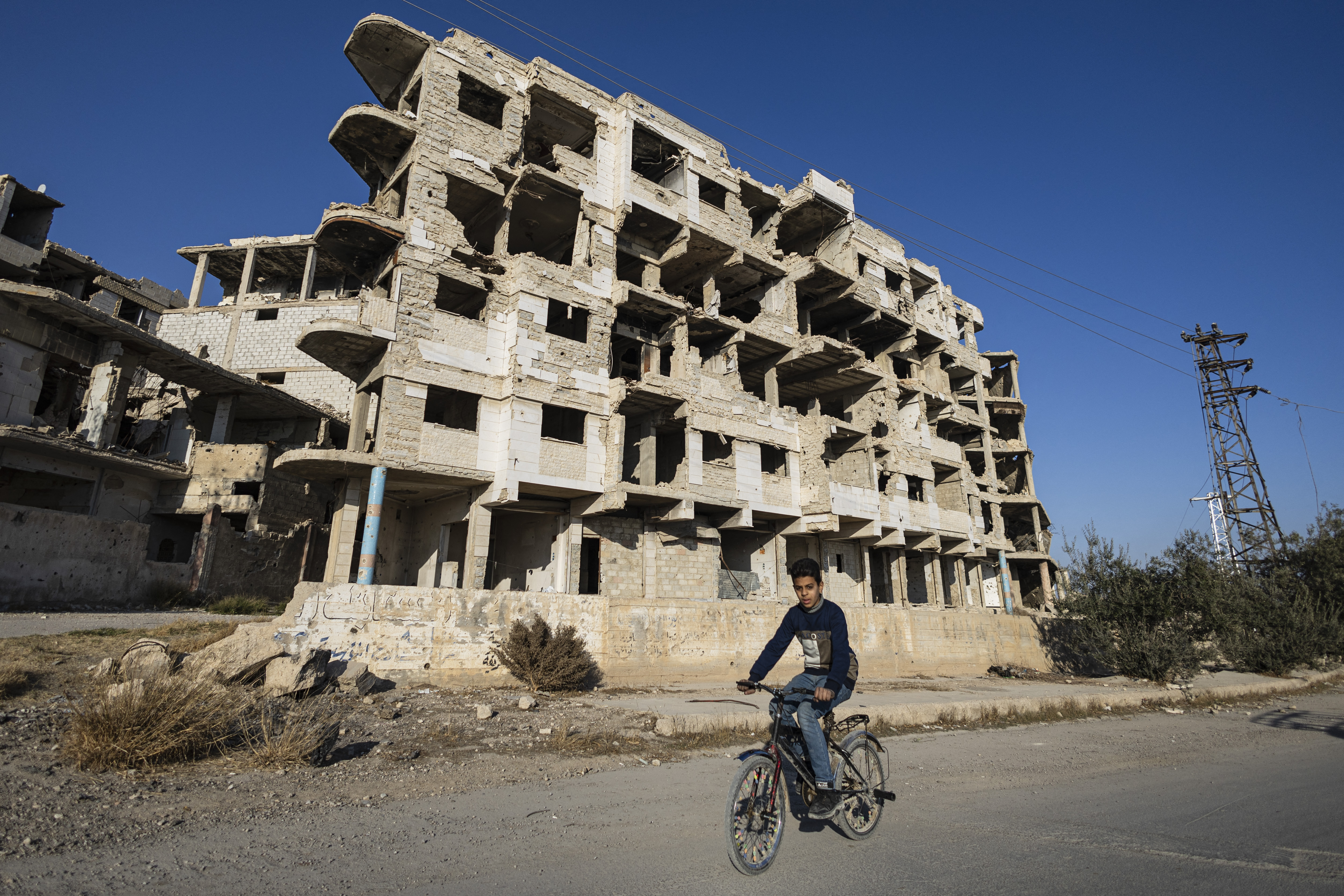 a boy rides a bicycle past a destroyed building