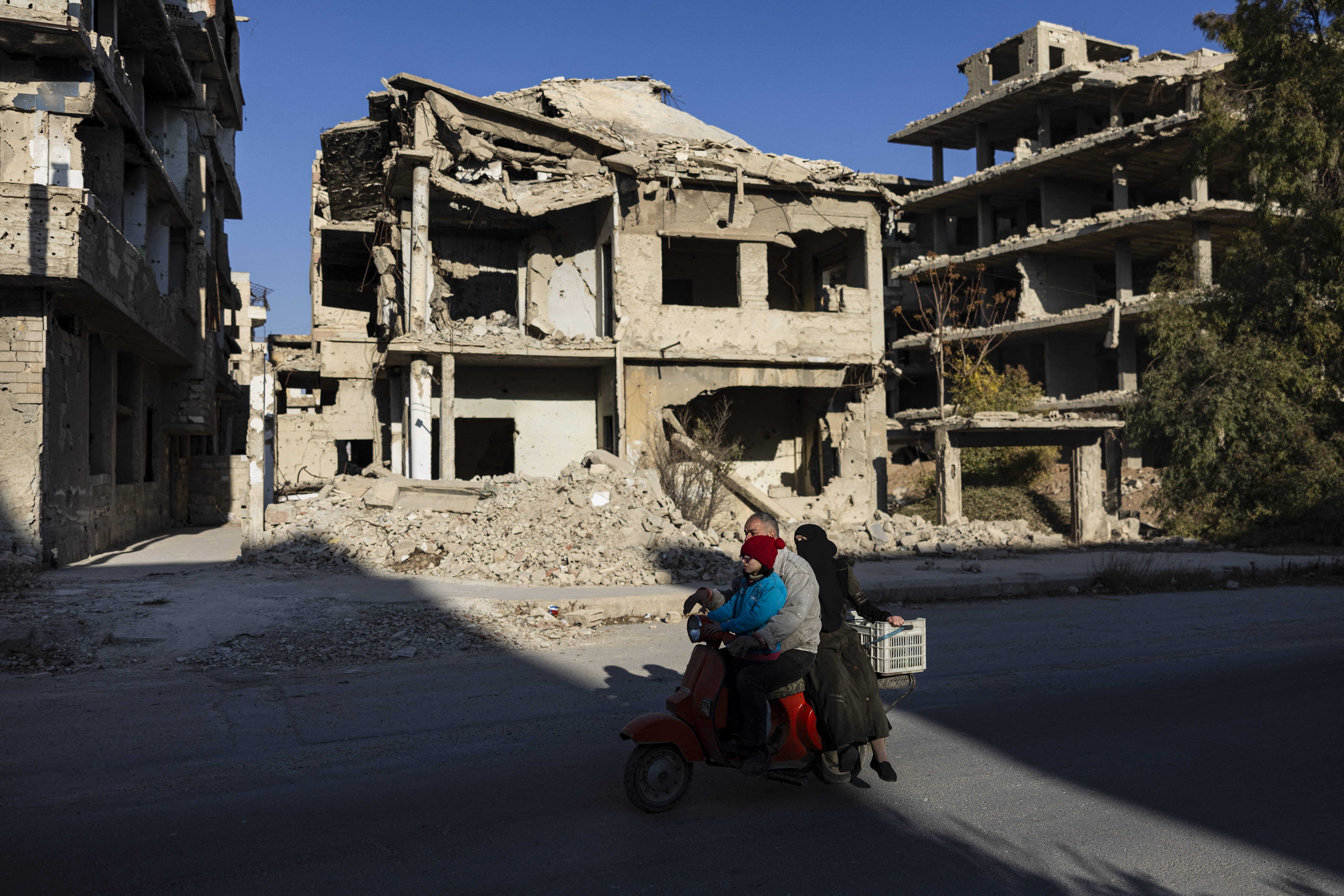 a family rides a motorcycle past a destroyed building