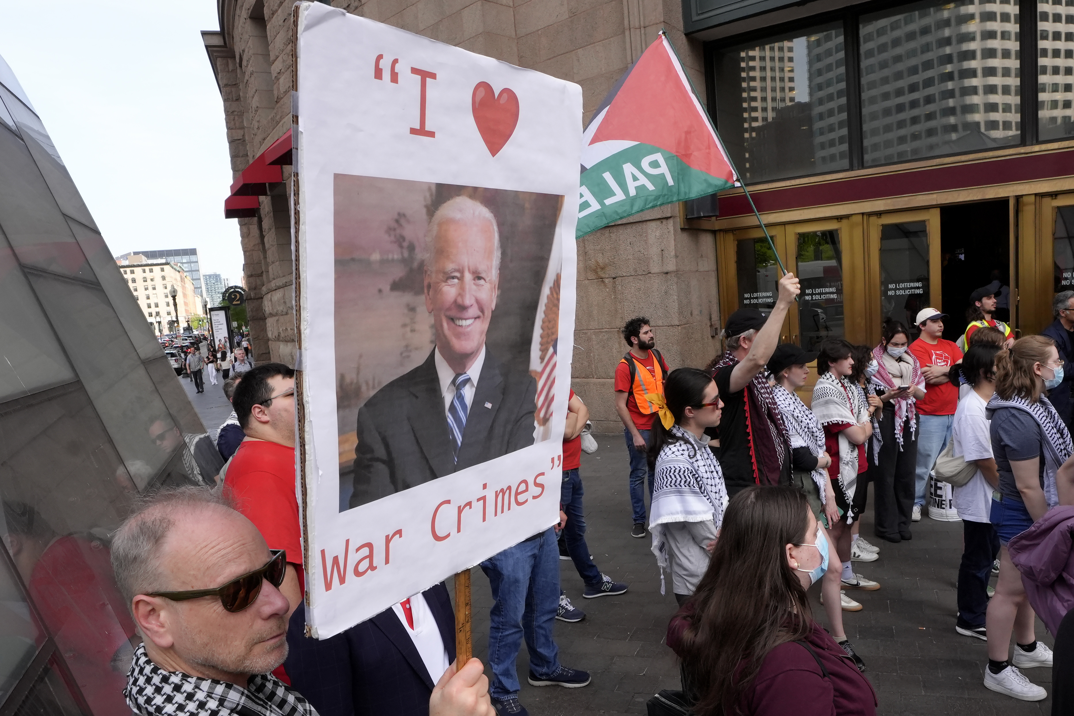 A protester in Boston holds up a picket sign that shows Joe Biden's face and the message, "I heart war crimes."
