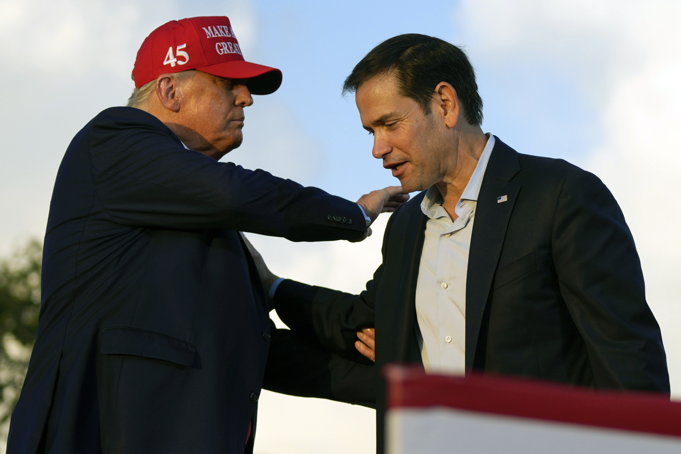 Donald Trump, wearing a red MAGA cap, reaches over to put a hand on Marco Rubio's shoulder