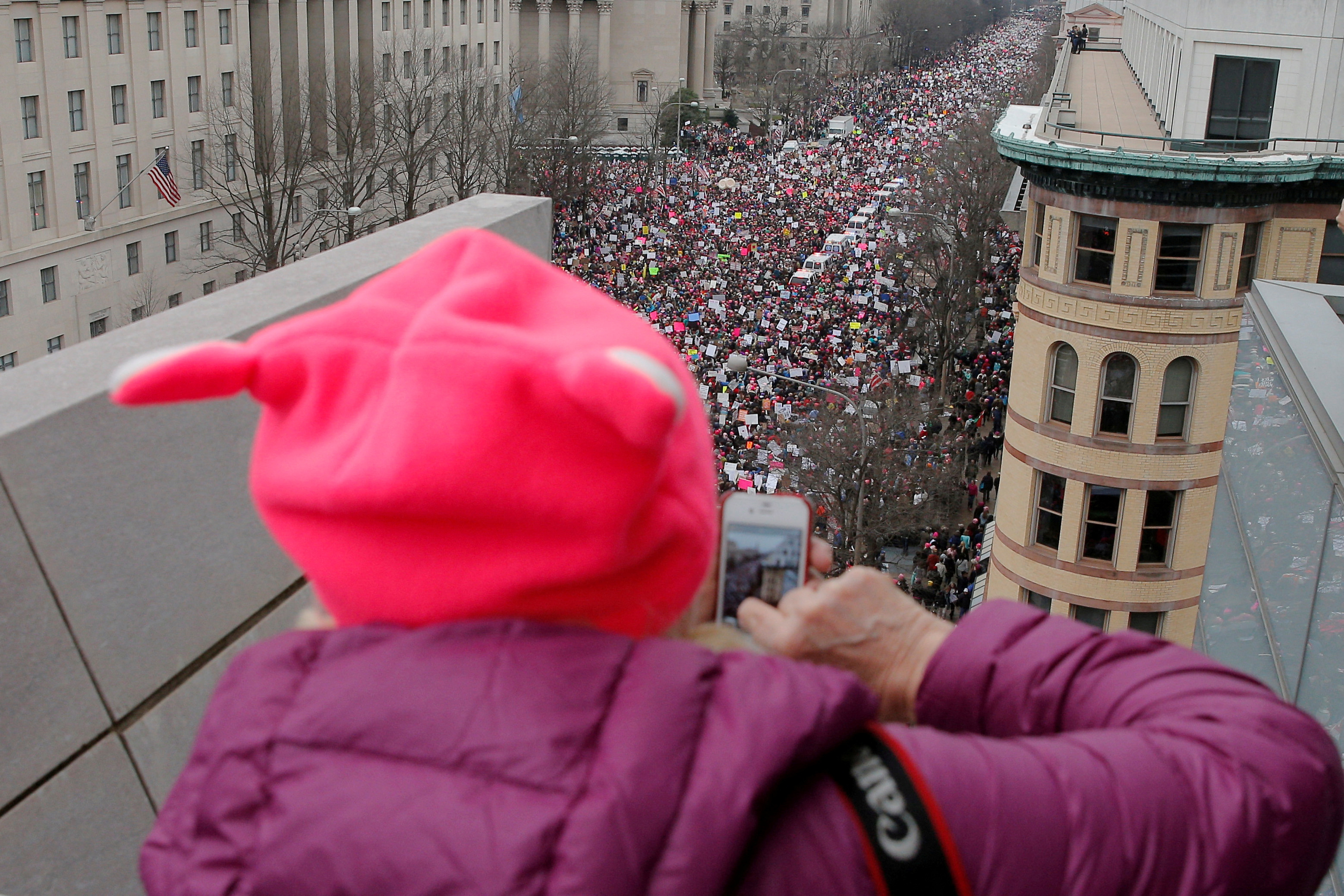 A person in a pink "pussy hat" takes a cellphone picture of the crowds below her building.